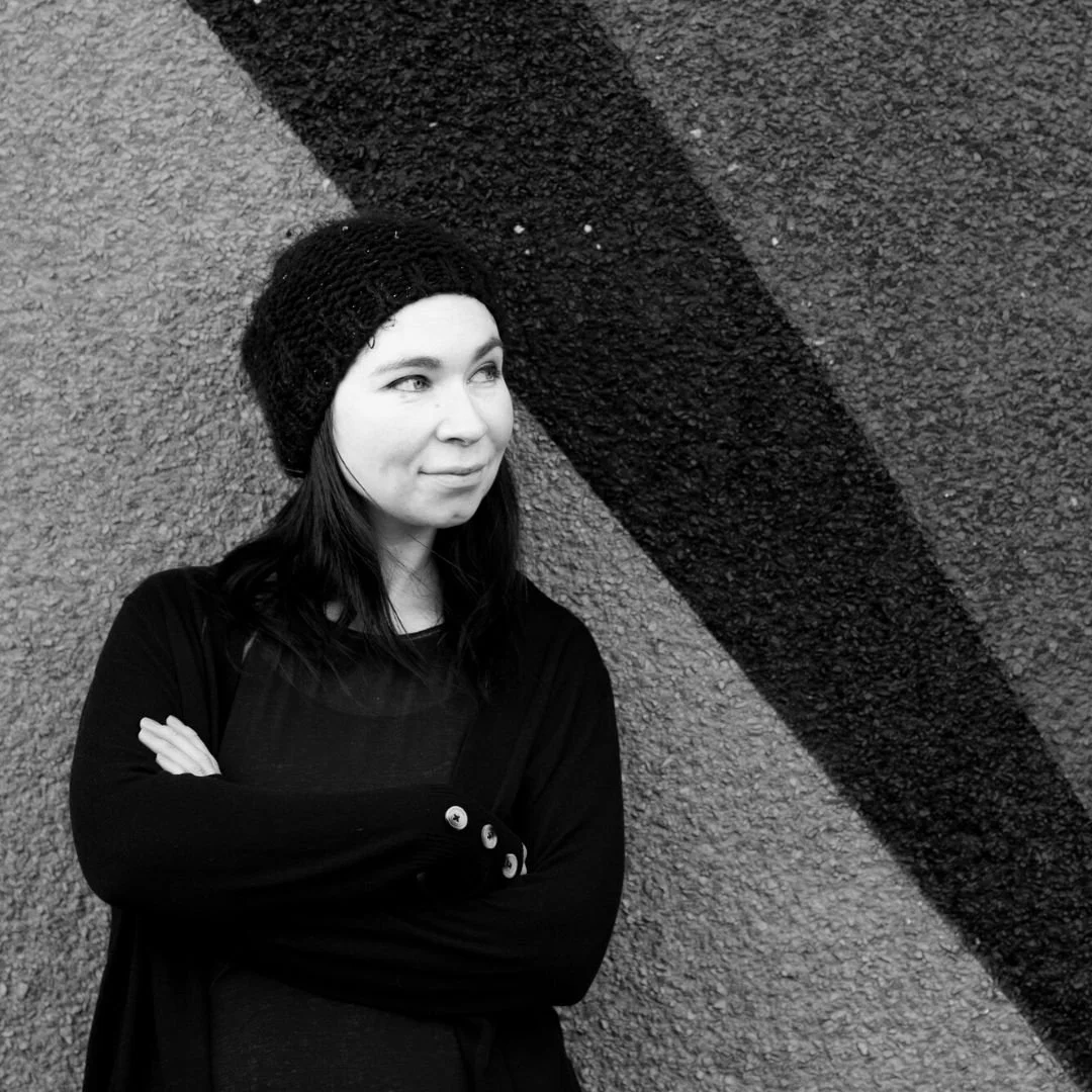A woman with long dark hair wearing a black knit hat and black long-sleeve top, standing with her arms crossed against a textured wall with a diagonal black stripe.
