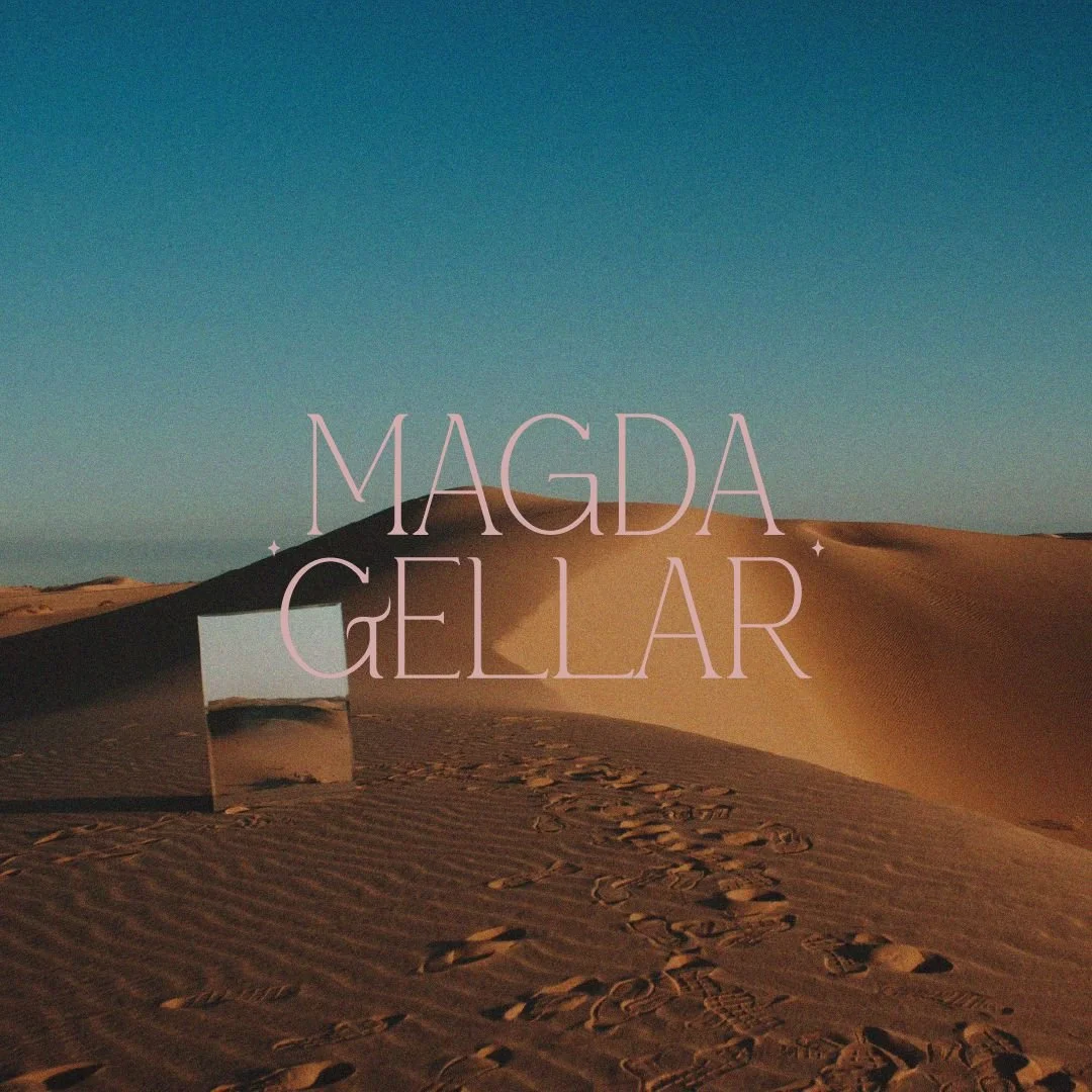 A mirror on a desert sand dune reflects the sky, with pink text overlay reading "Magda Gellar" against a clear blue sky.