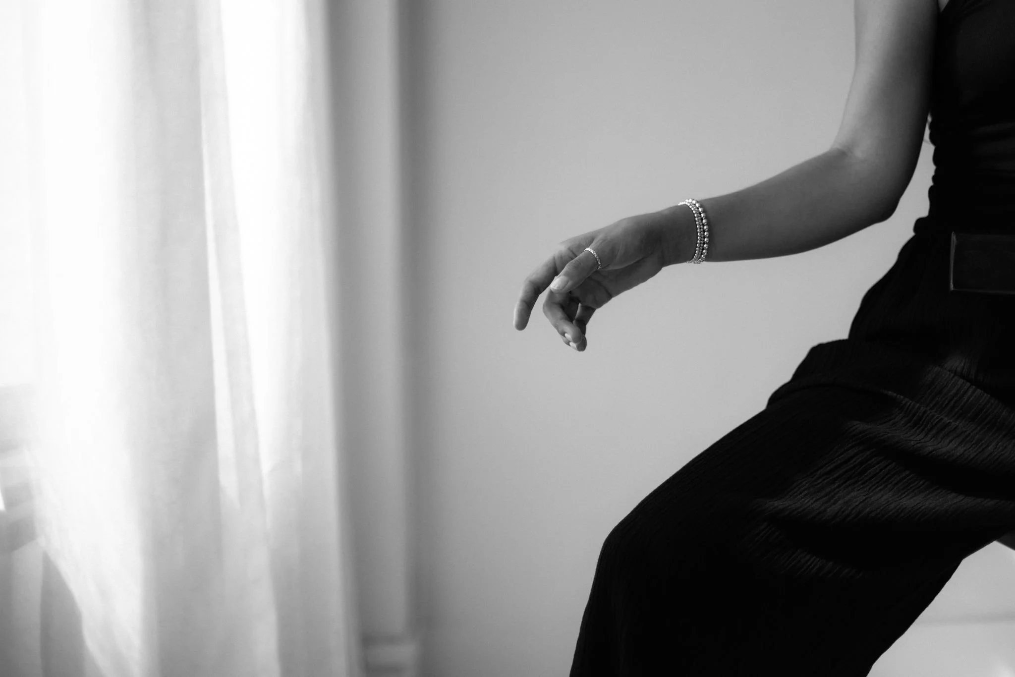 Black and white photo of a woman's arm and hand, with jewelry, resting on her knee, with a blurred curtain in the background.