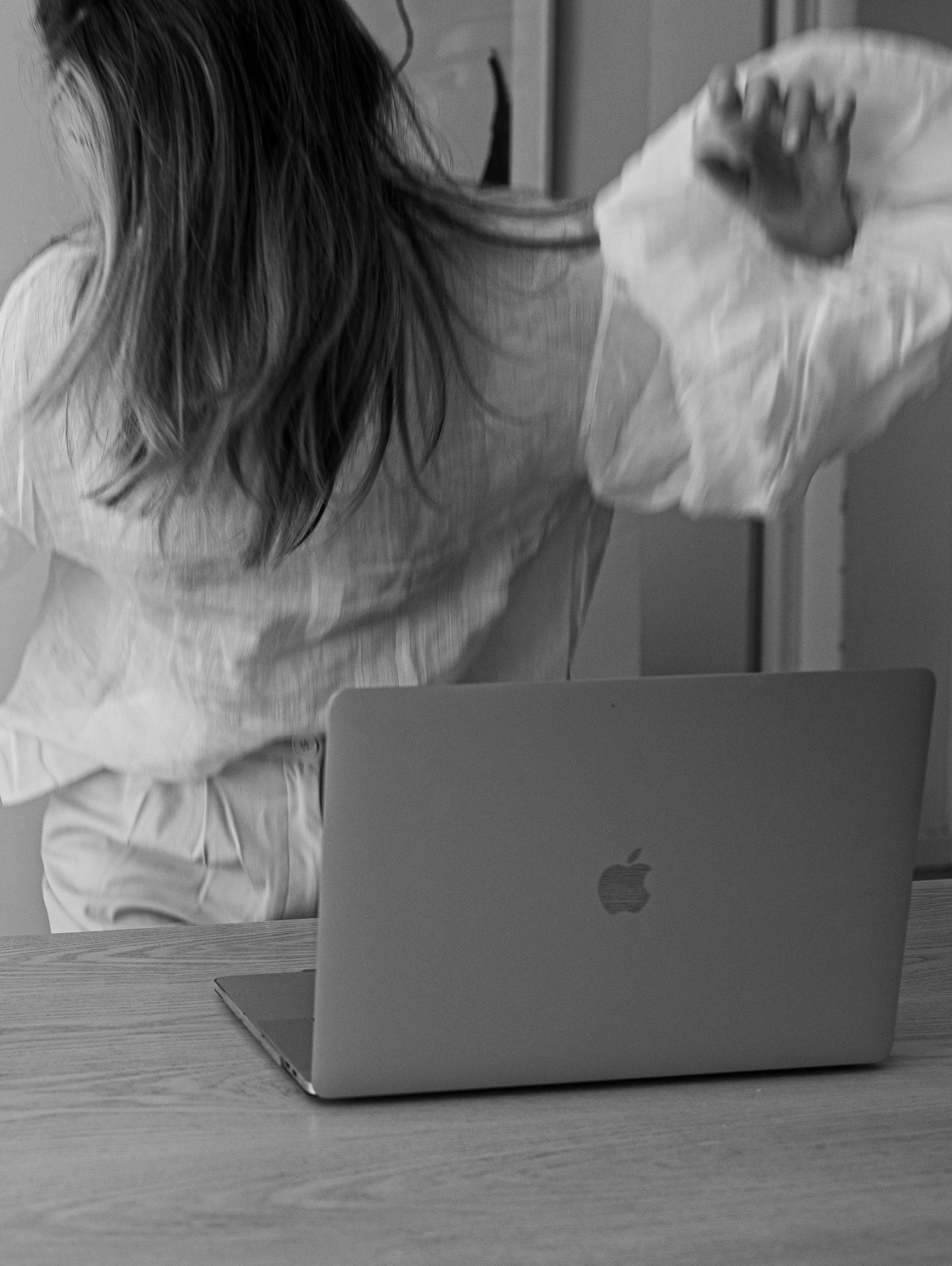 Person with long hair wearing a loose shirt, leaning over a table with a closed Apple MacBook laptop, reaching out with one hand, in a grayscale photo.
