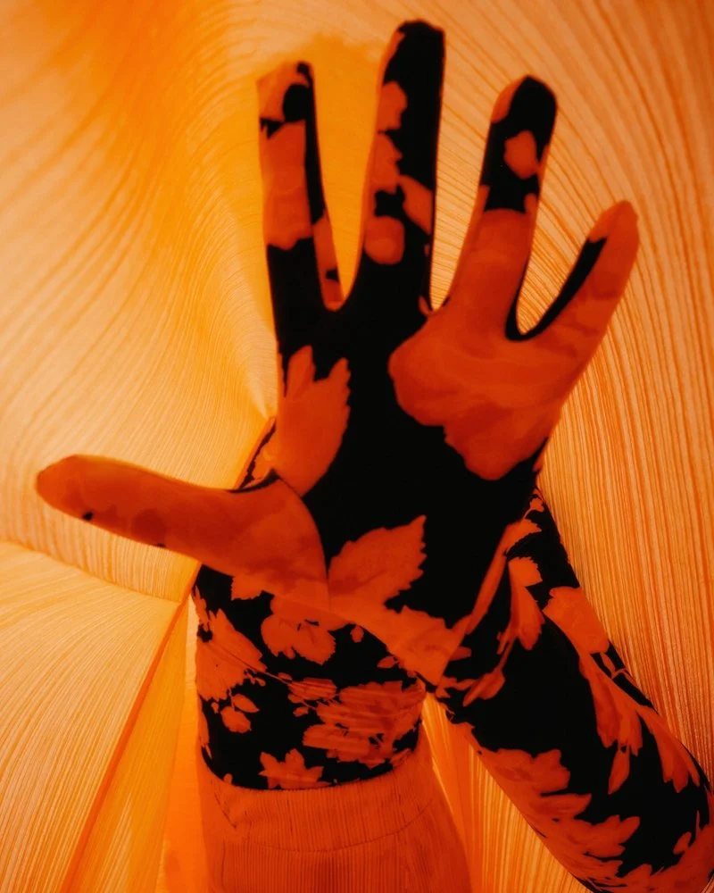 A person's hand and arm covered with a black and orange floral patterned long sleeve shirt or glove, reaching inside a bright orange paper lantern with vertical ridges.
