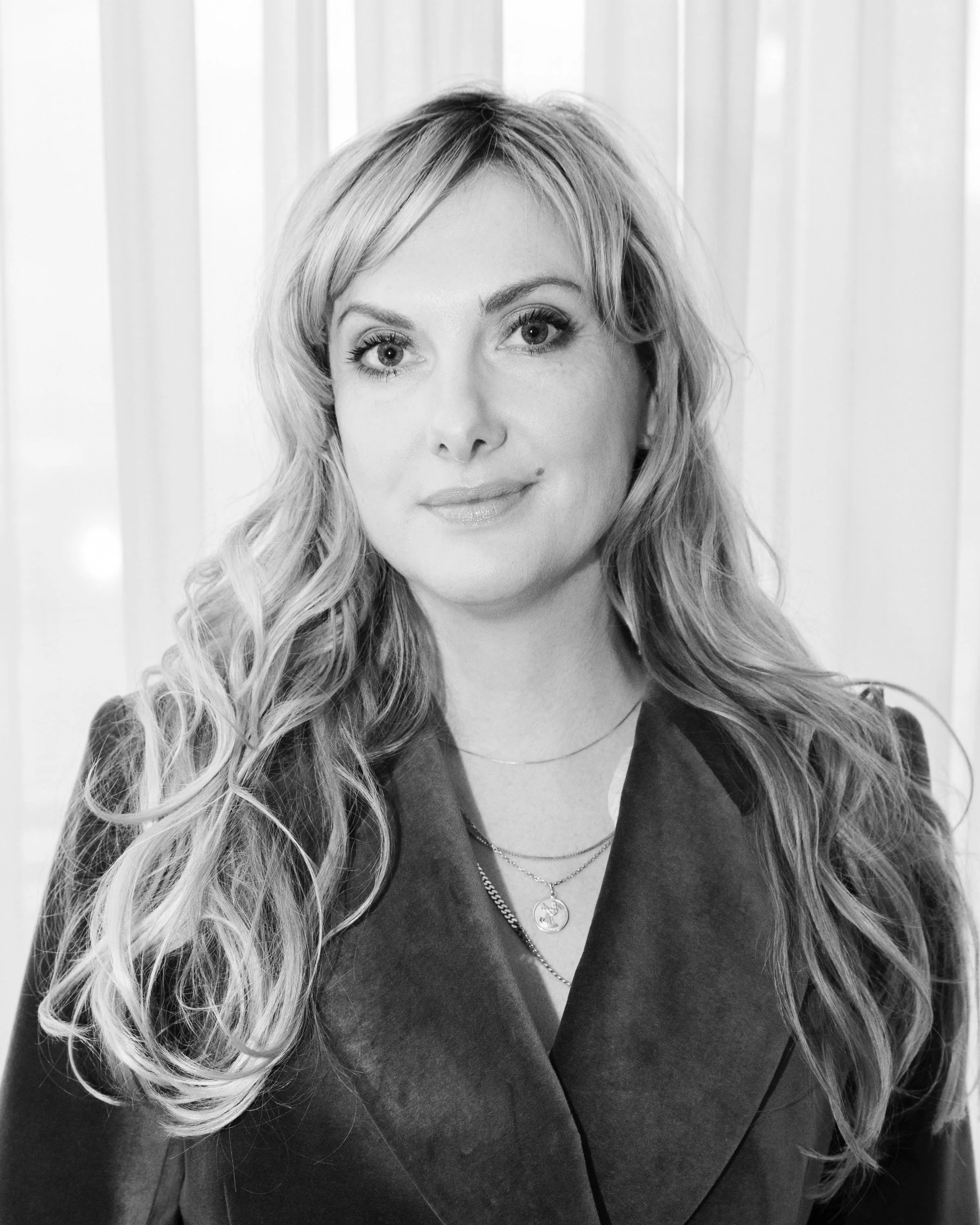 A black-and-white portrait of Nina Knag with long, wavy hair, wearing a dark blazer and layered necklaces, standing in front of curtains.