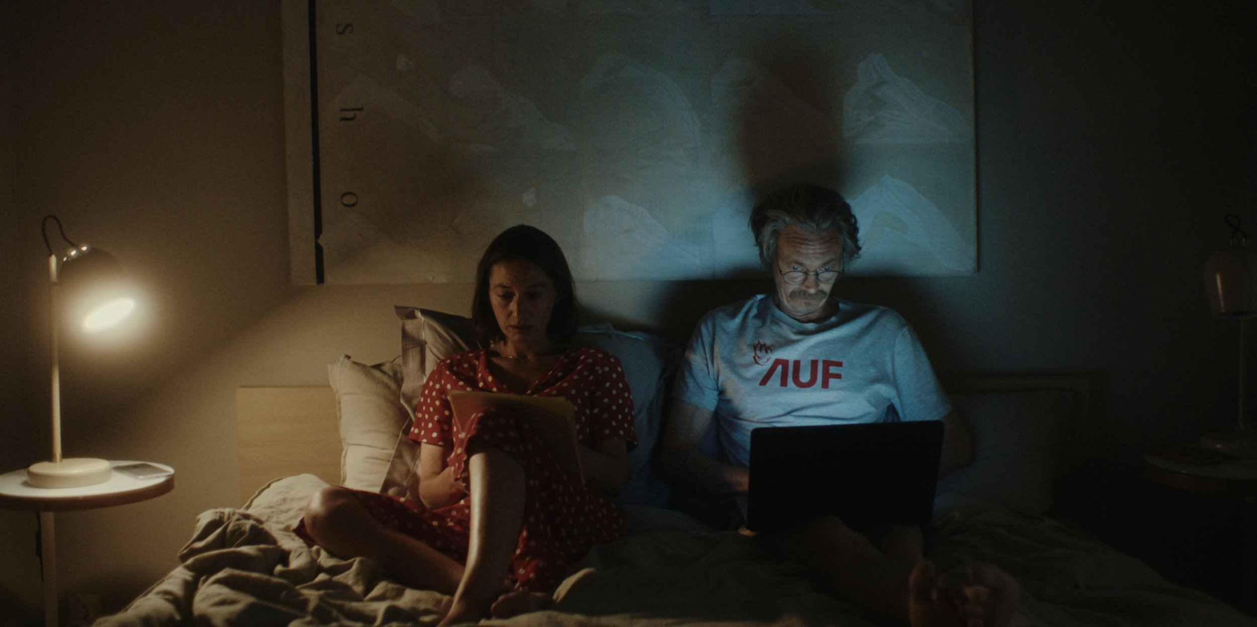 A woman in a red polka dot dress and a man in a gray T-shirt with red and black text are sitting on a bed at night, using a laptop and a tablet. The room is dimly lit by two small lamps on nightstands.