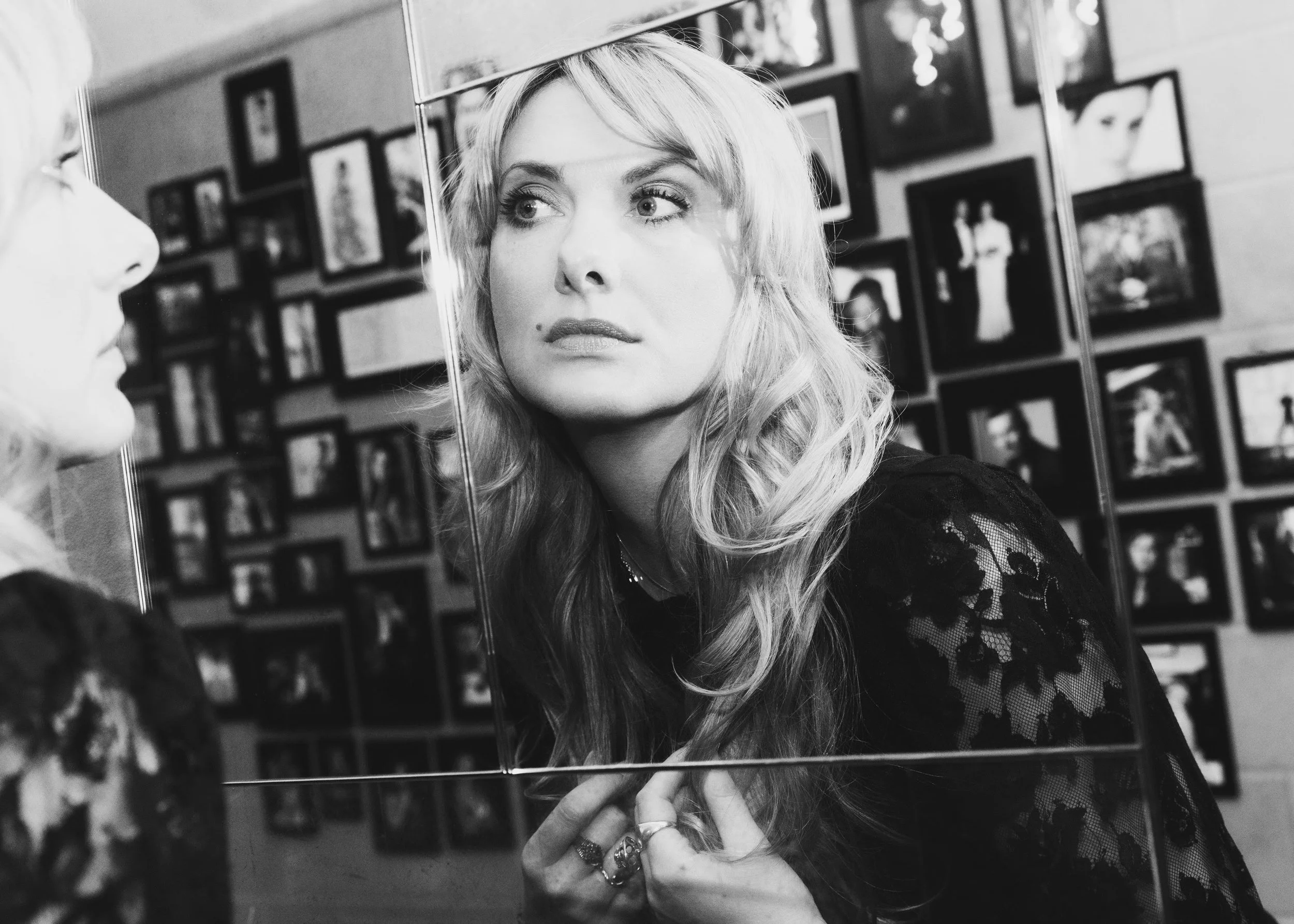 Nina Knag with wavy blonde hair looking at her reflection in a mirror, with multiple framed photographs on the wall behind her, in black and white.