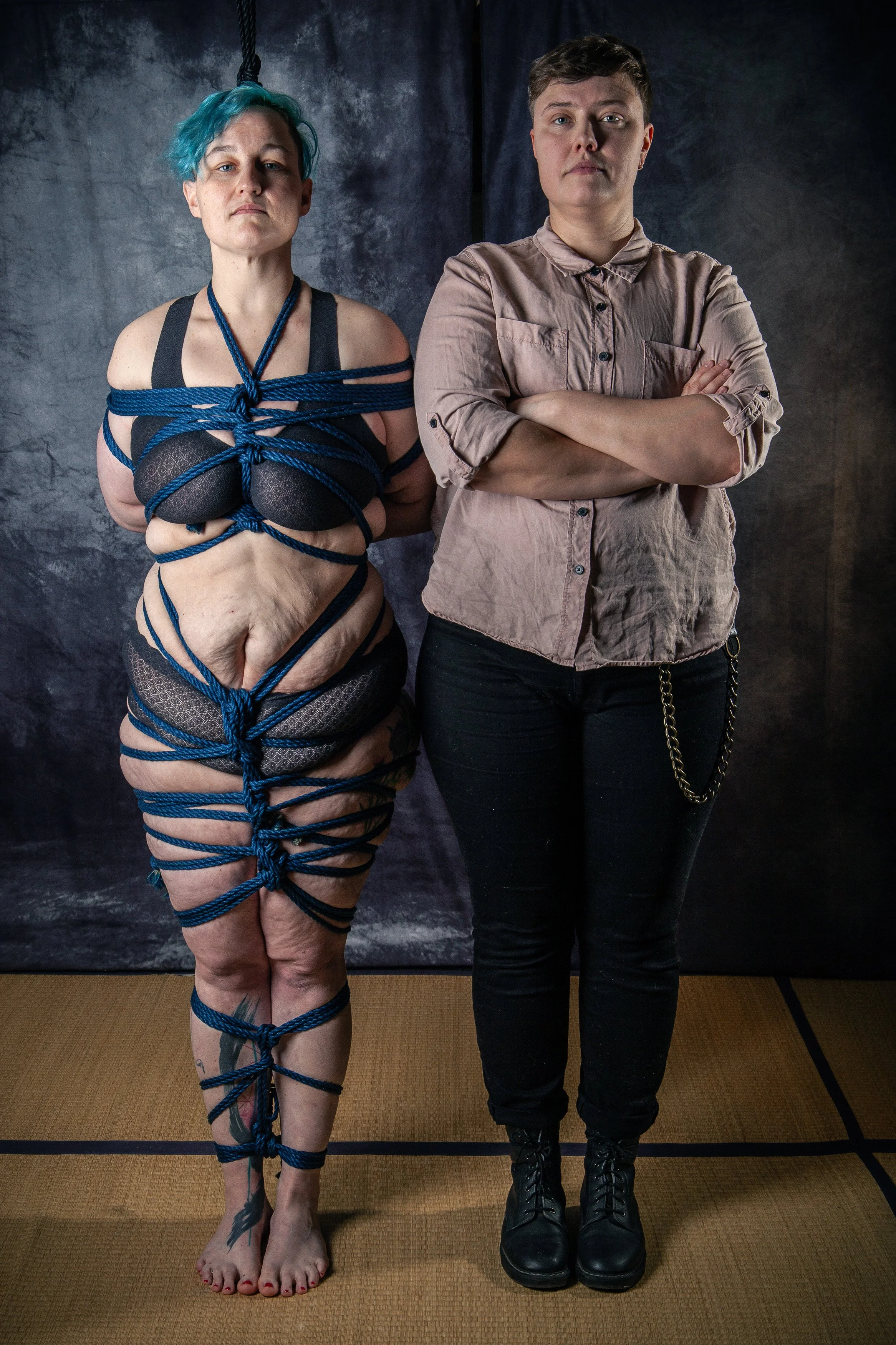 A person wrapped tightly in dark blue ropes with a security knot, standing next to another person with arms crossed. The background is a dark textured curtain, and they are on a tatami mat floor.