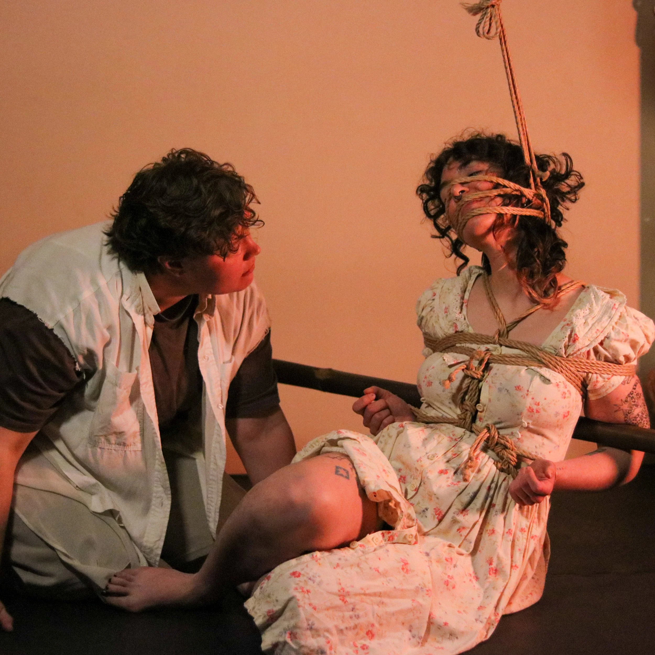 A woman tied with ropes around her chest and neck, seated on a bed, with a rope hanging from above near her face. A man kneels next to her, holding her foot, looking at her.