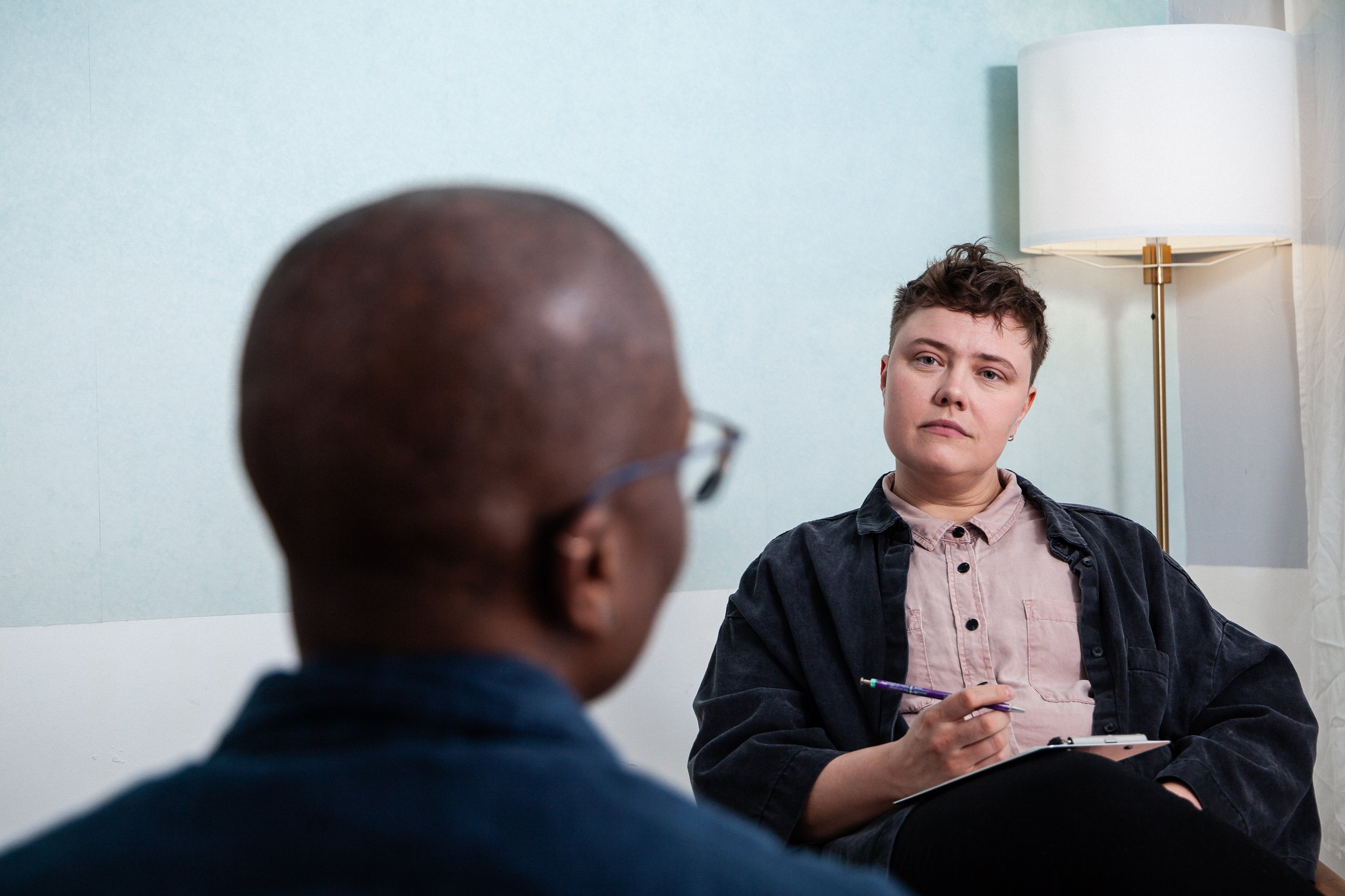 A woman with short hair sitting in a therapy session, holding a notebook and pen, looking thoughtfully at a psychologist with glasses, in a room with a lamp and light blue wall.