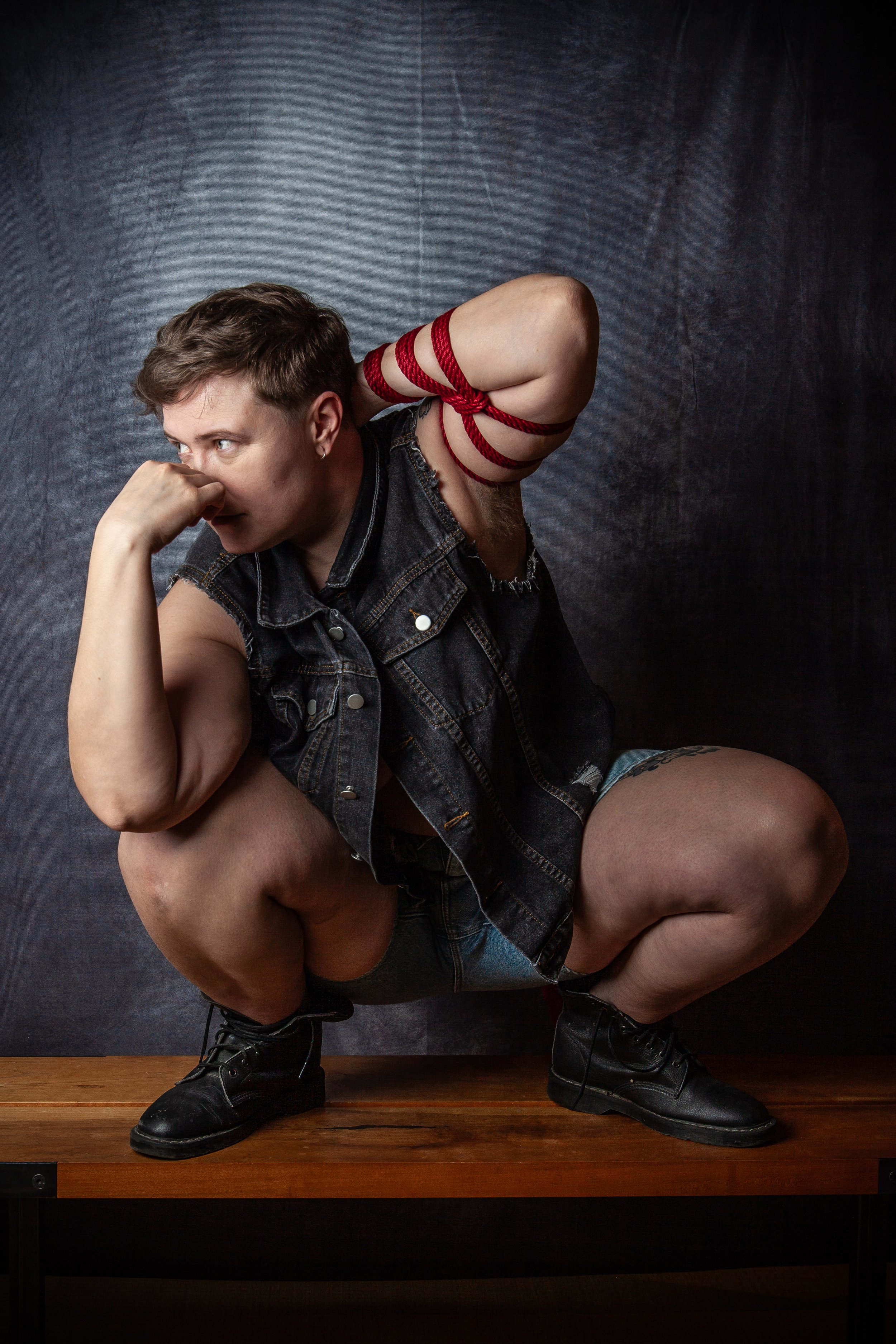 Person with short hair wearing sleeveless denim vest squatting on a wooden surface against a dark textured background, with red rope wrapped around their right arm and leg, and black boots.