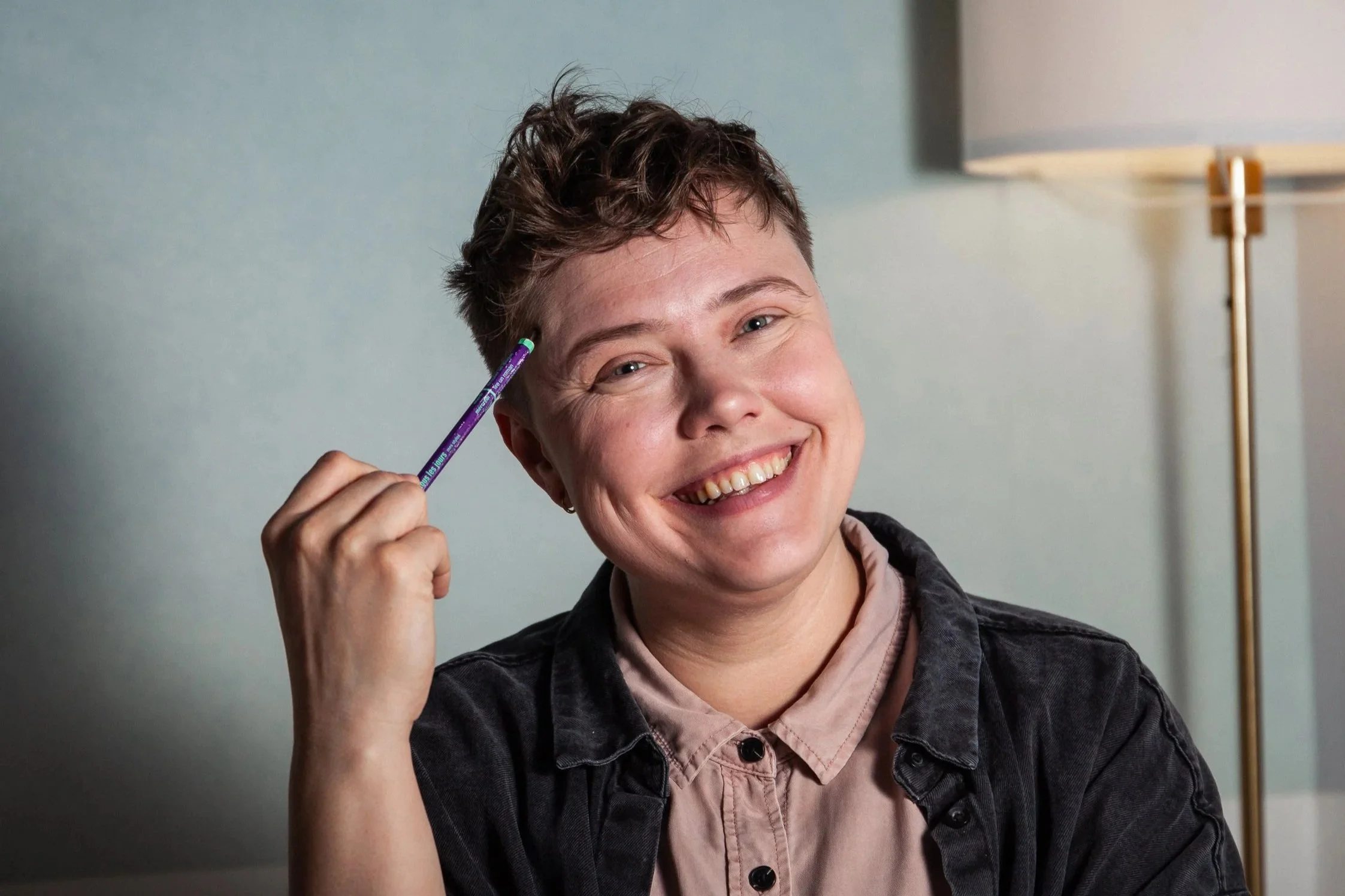 A person with short, curly brown hair smiling, holding a purple eyeliner pencil near their temple, wearing a light pink shirt and dark jacket, in front of a gray wall and standing next to a floor lamp.
