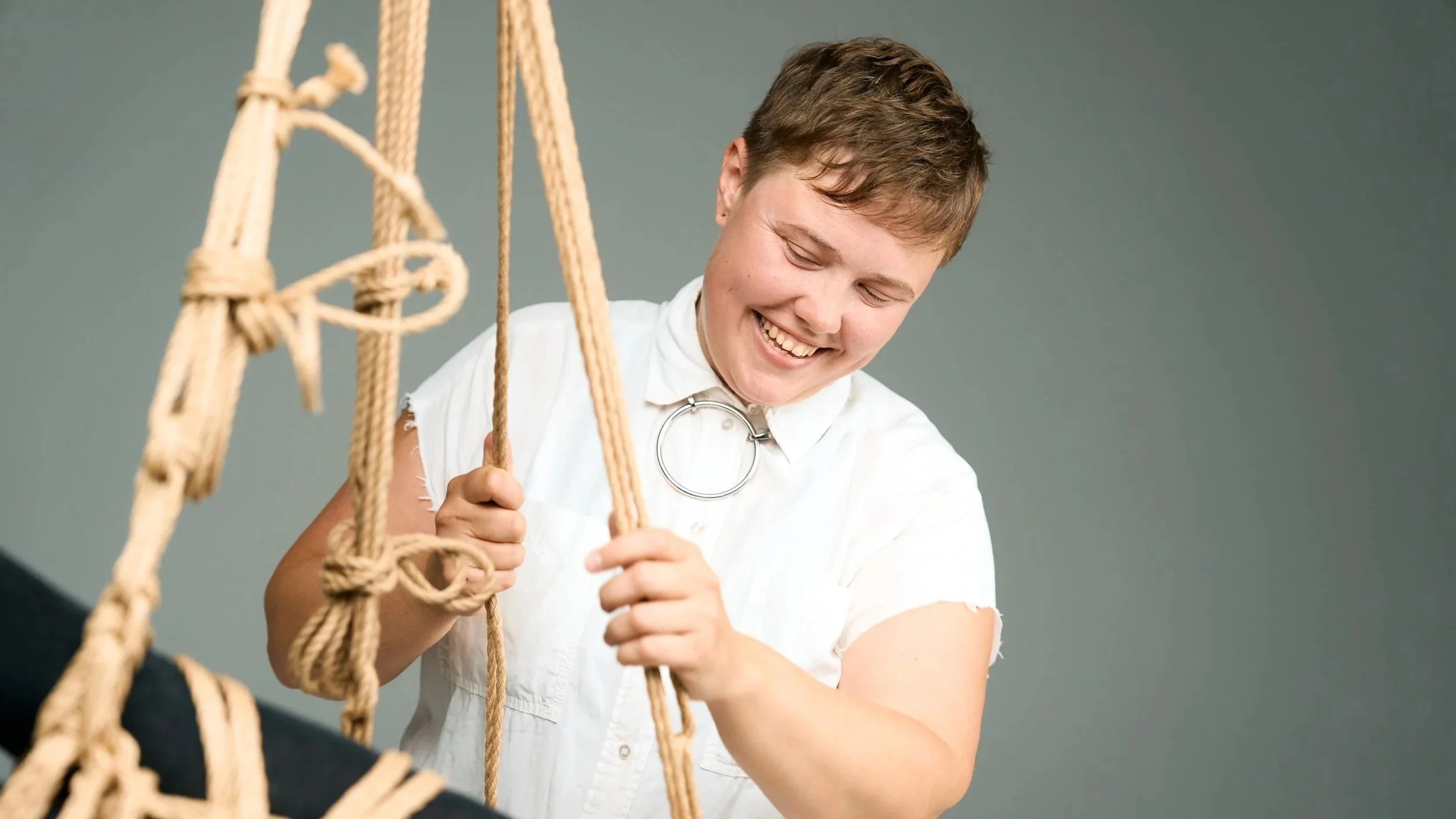 Person smiling and climbing on a rope ladder against a gray background.