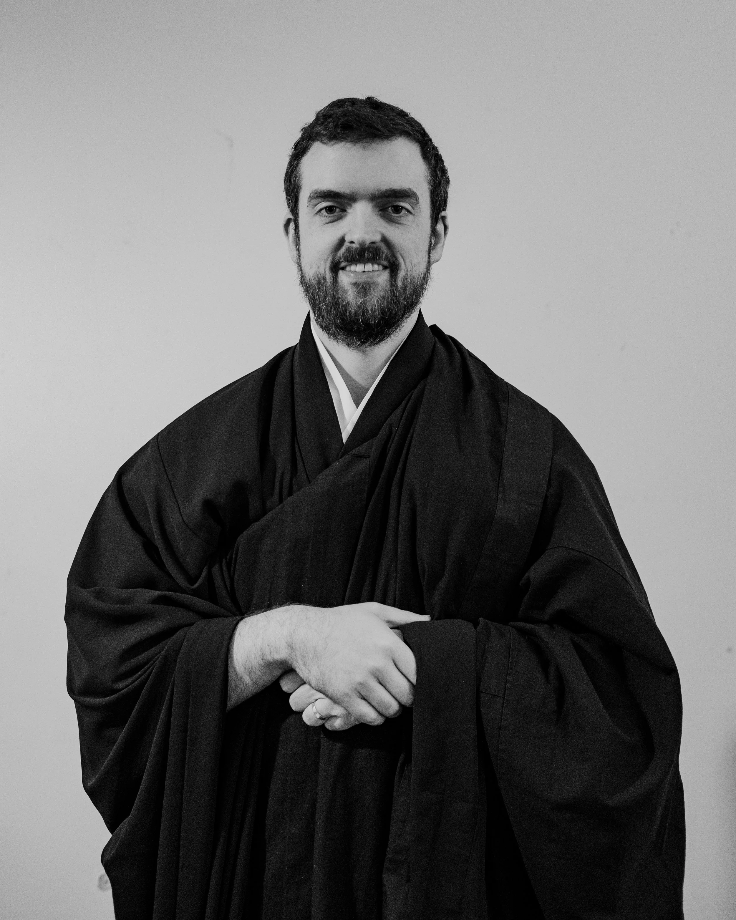 A man dressed in traditional Buddhist monk robes, standing indoors with a neutral expression.