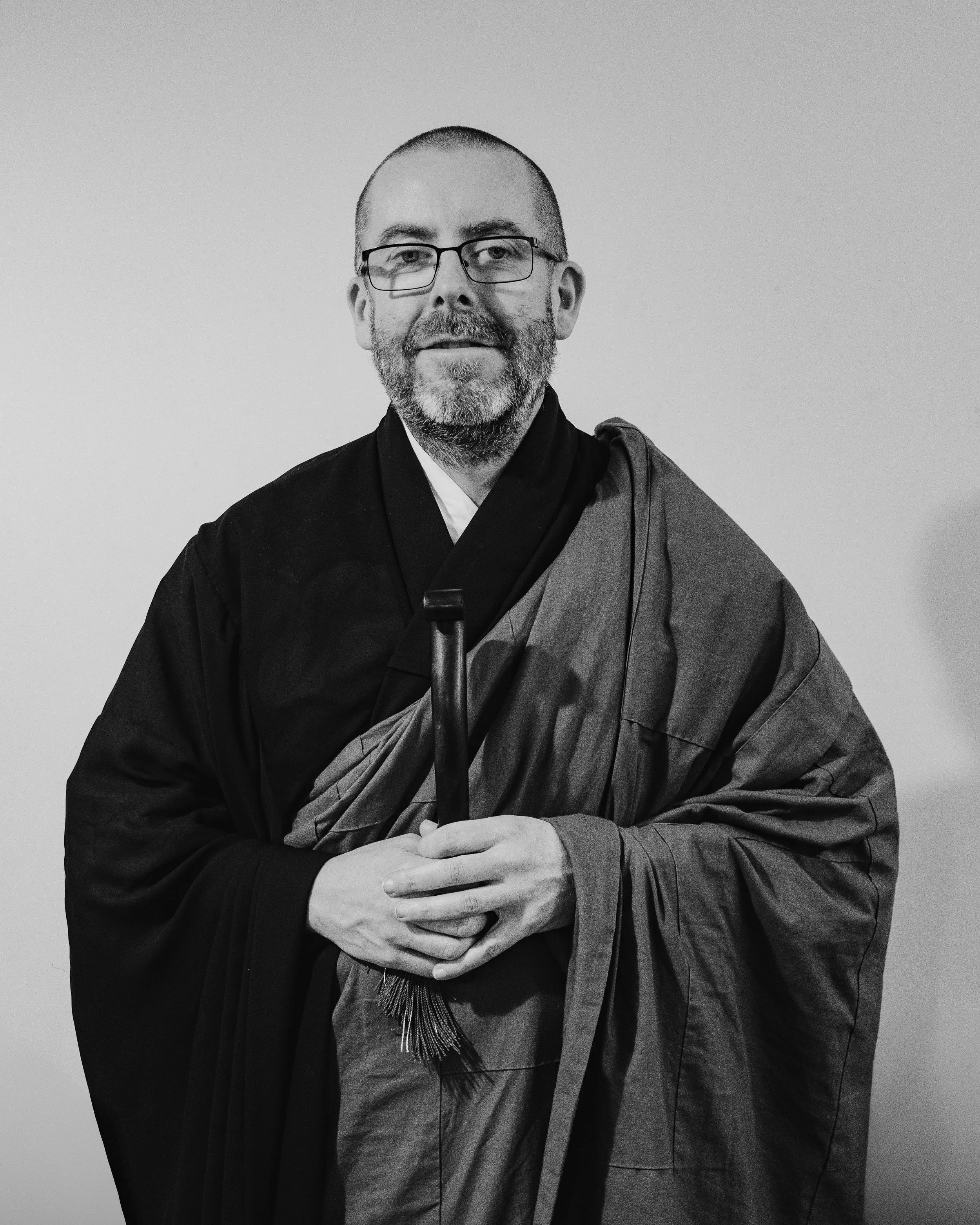 A man with glasses and a beard smiling and laughing while sitting at a table with a ceramic cup, in front of a window with raindrops, in black and white.