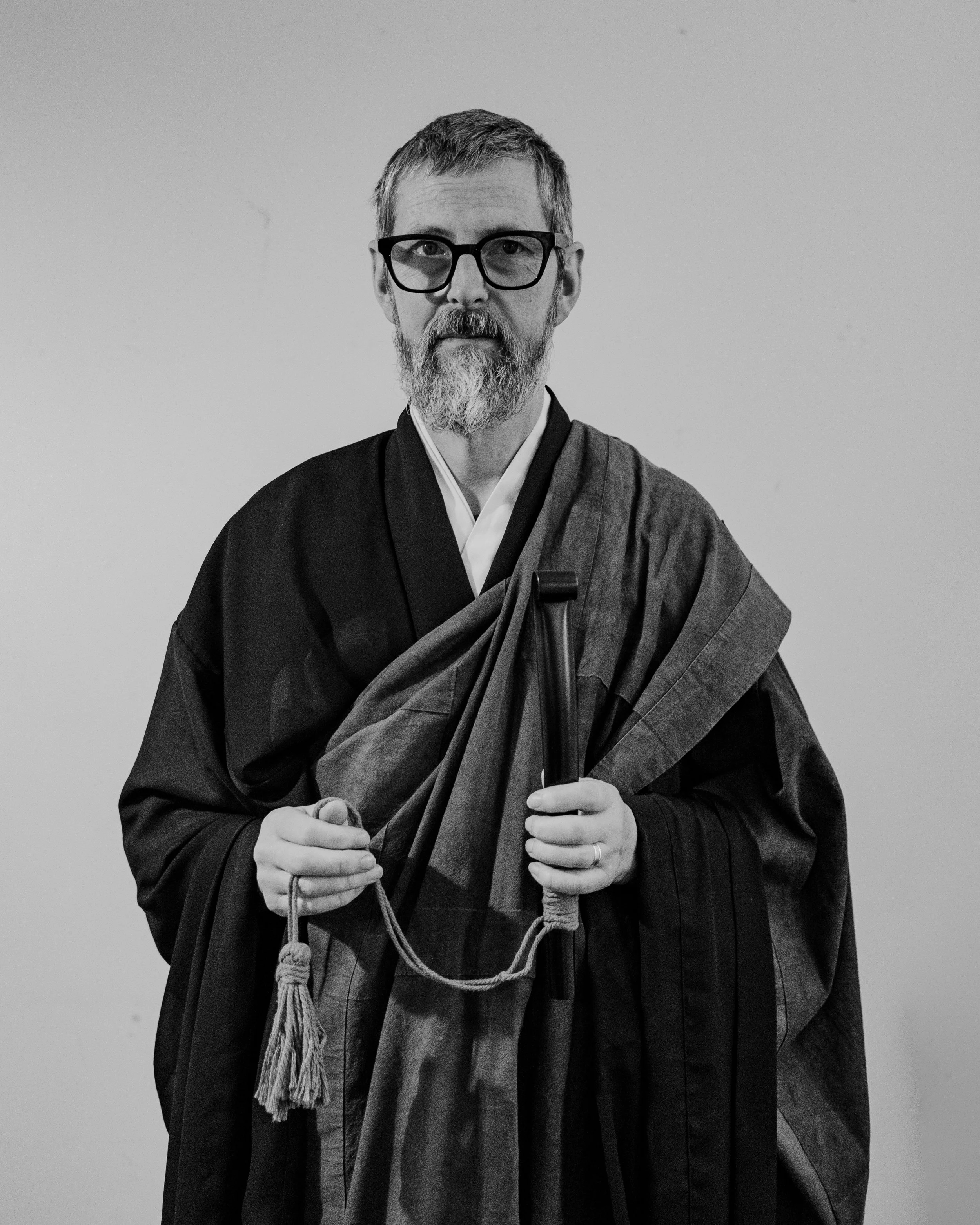 Black and white photo of a monk sitting at a table in a cozy room with books and photos on shelves, and flags hanging near a window.