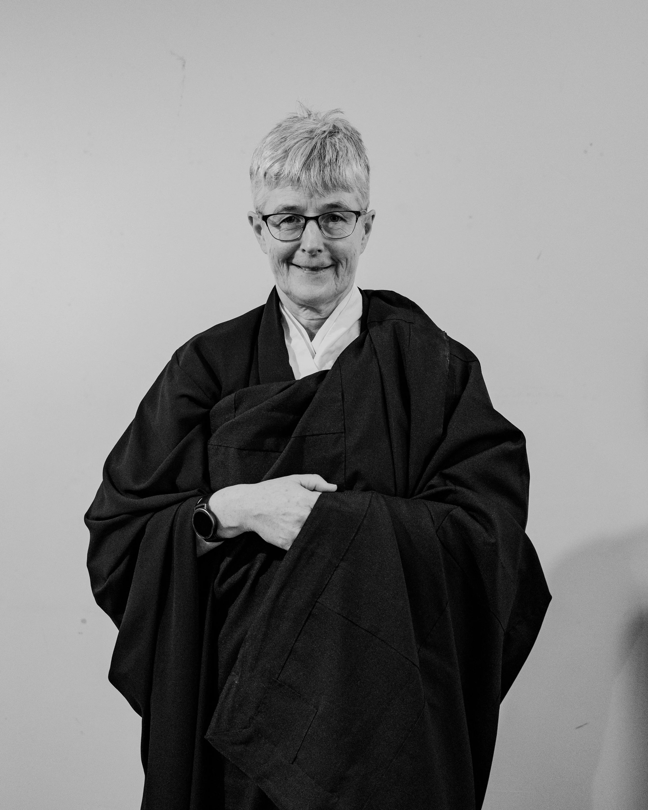 A person dressed in a graduation gown and cap, smiling outdoors with a large bush in the background.