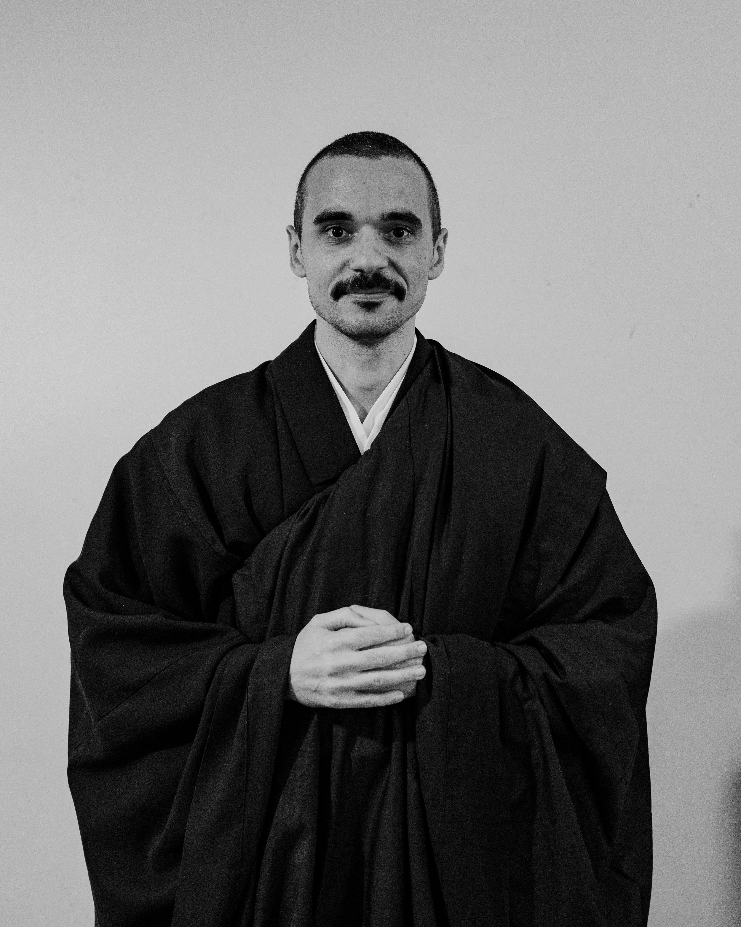 Black and white photo of a bald man in traditional monk robes with hands pressed together in a prayer pose standing in a room with arched doorway and framed picture in background.