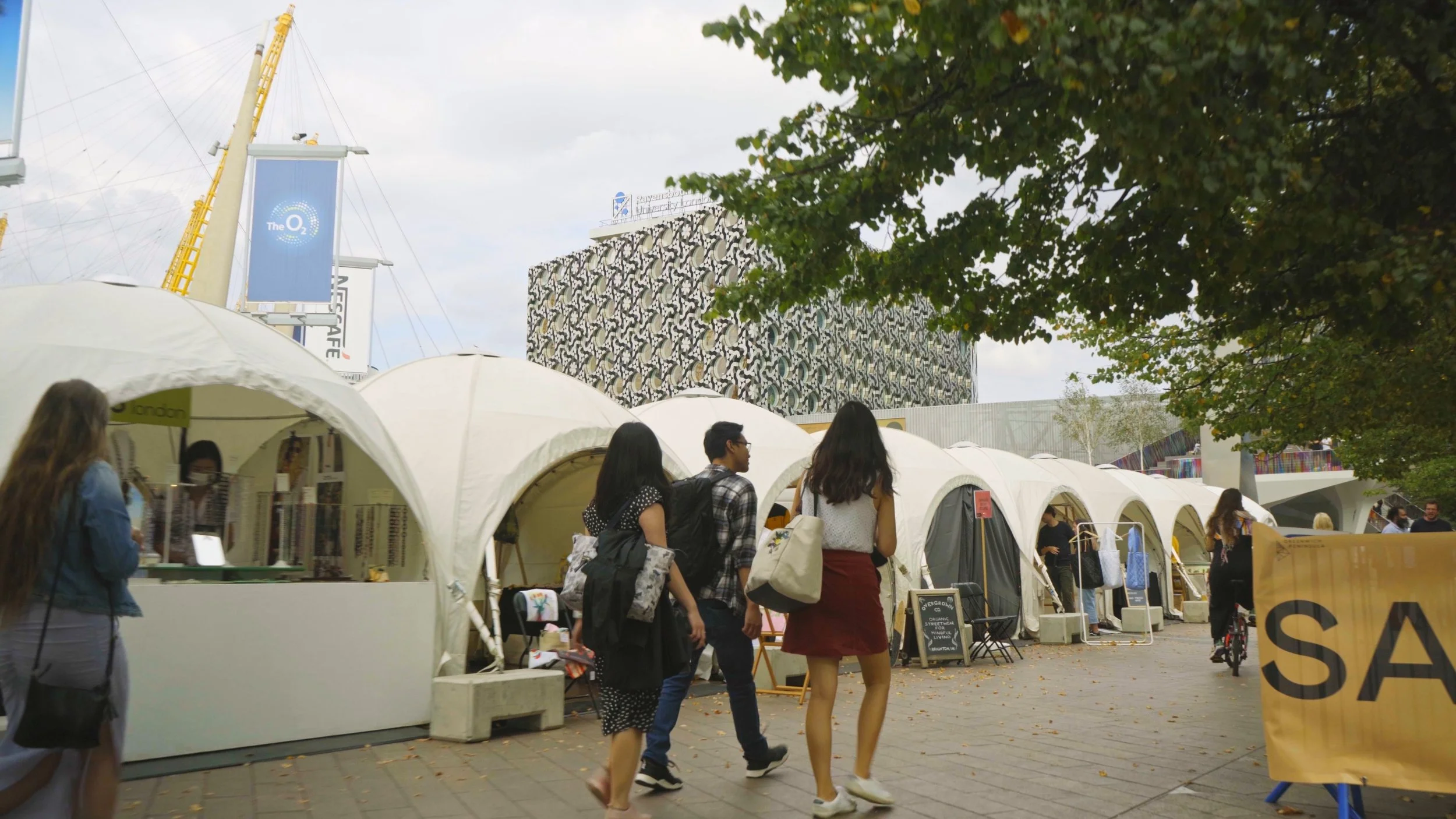People walking past white tents at an outdoor event in a city, with a modern building and trees in the background.