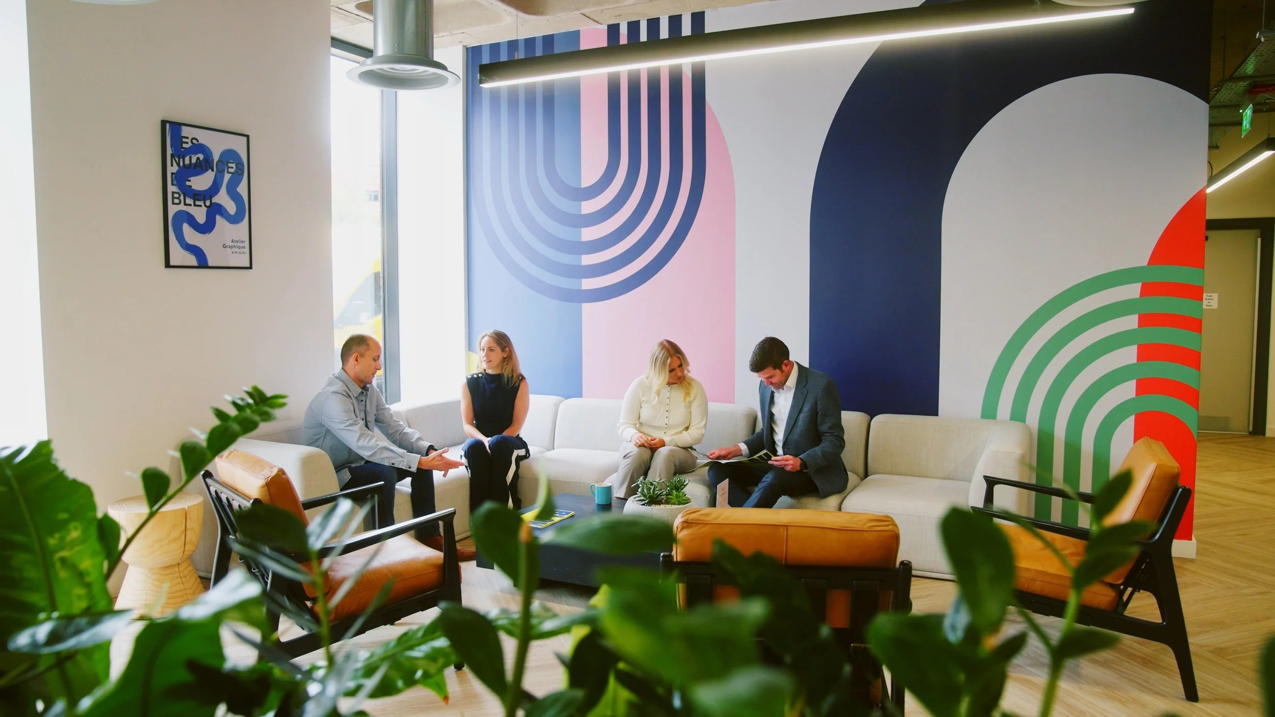 A group of five people sitting on a white sofa in a modern office lounge area with colorful abstract wall art, plants, and chairs.