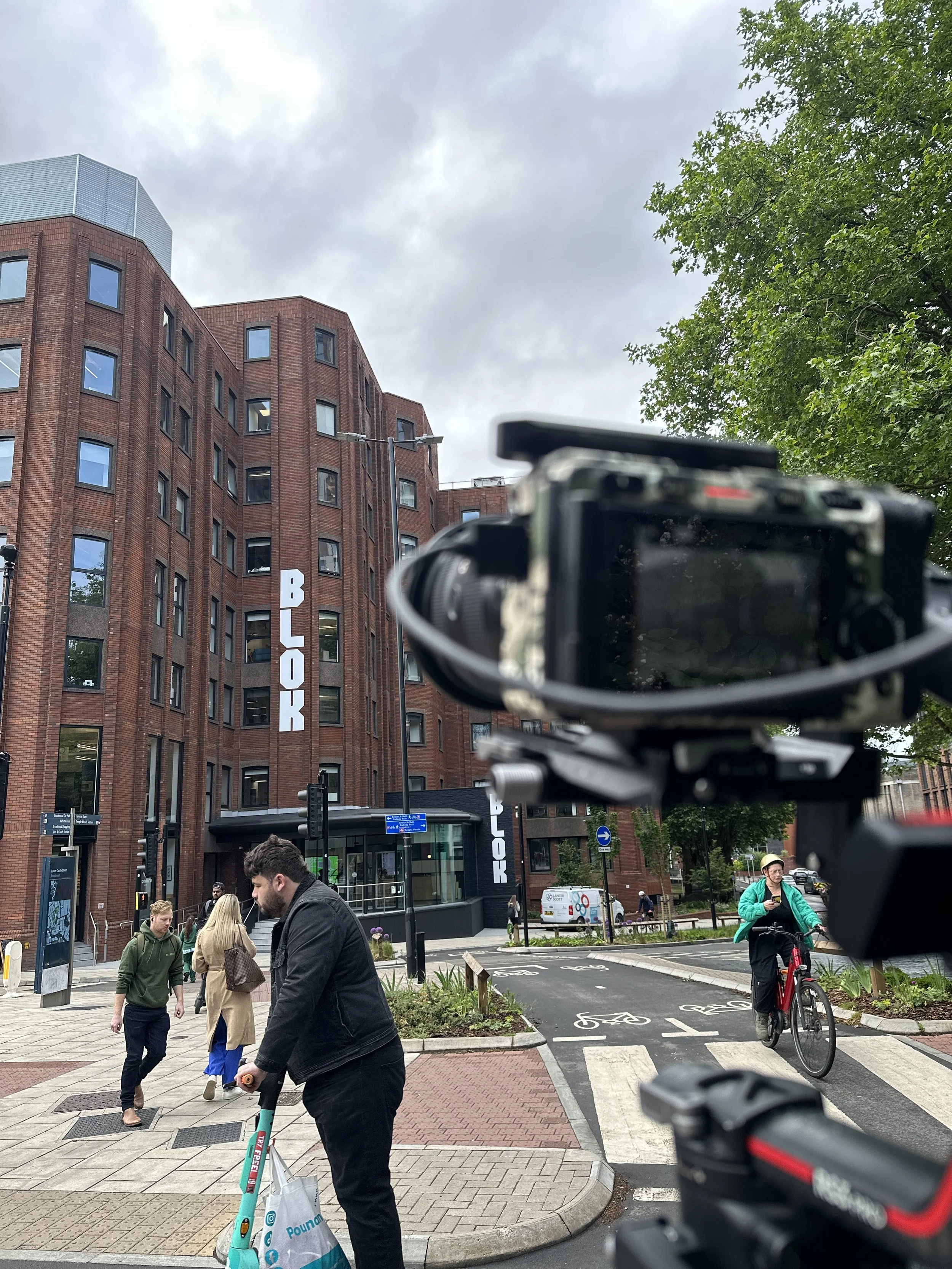 A city street scene with a brick building labeled 'BLOCK' in the background, people walking and riding bikes, and a camera in the foreground capturing the view.