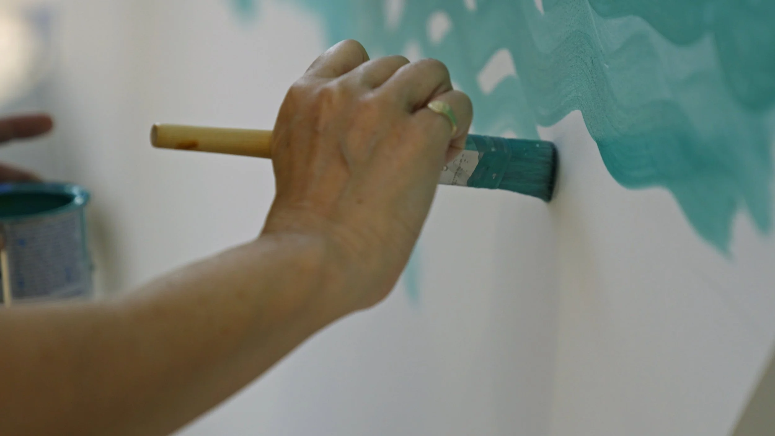 A person using a paintbrush to paint teal-colored shapes on a wall with a small open can of teal paint nearby.