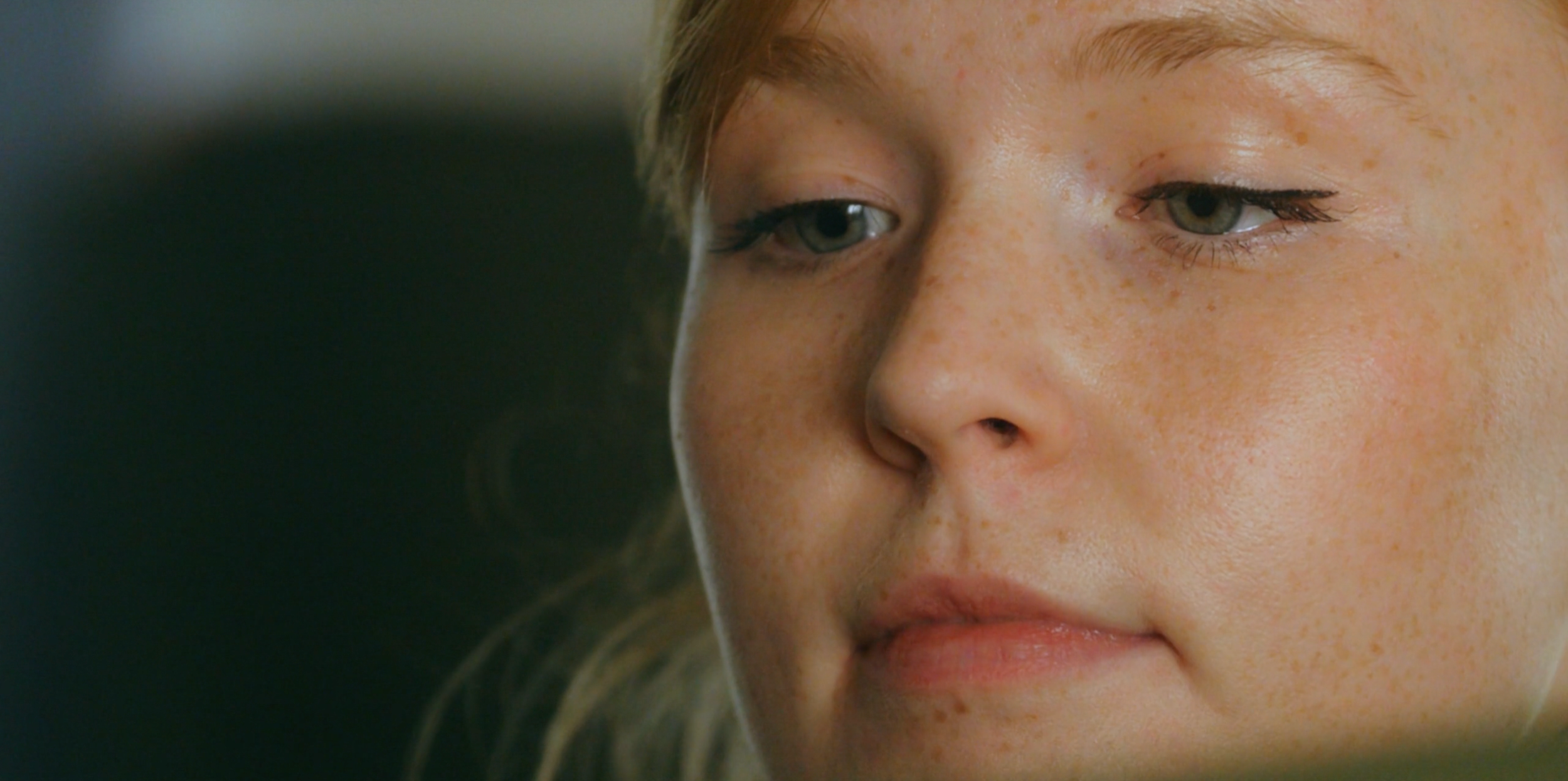 Close-up of a young woman's face with light skin and freckles, looking slightly downward with a neutral expression.