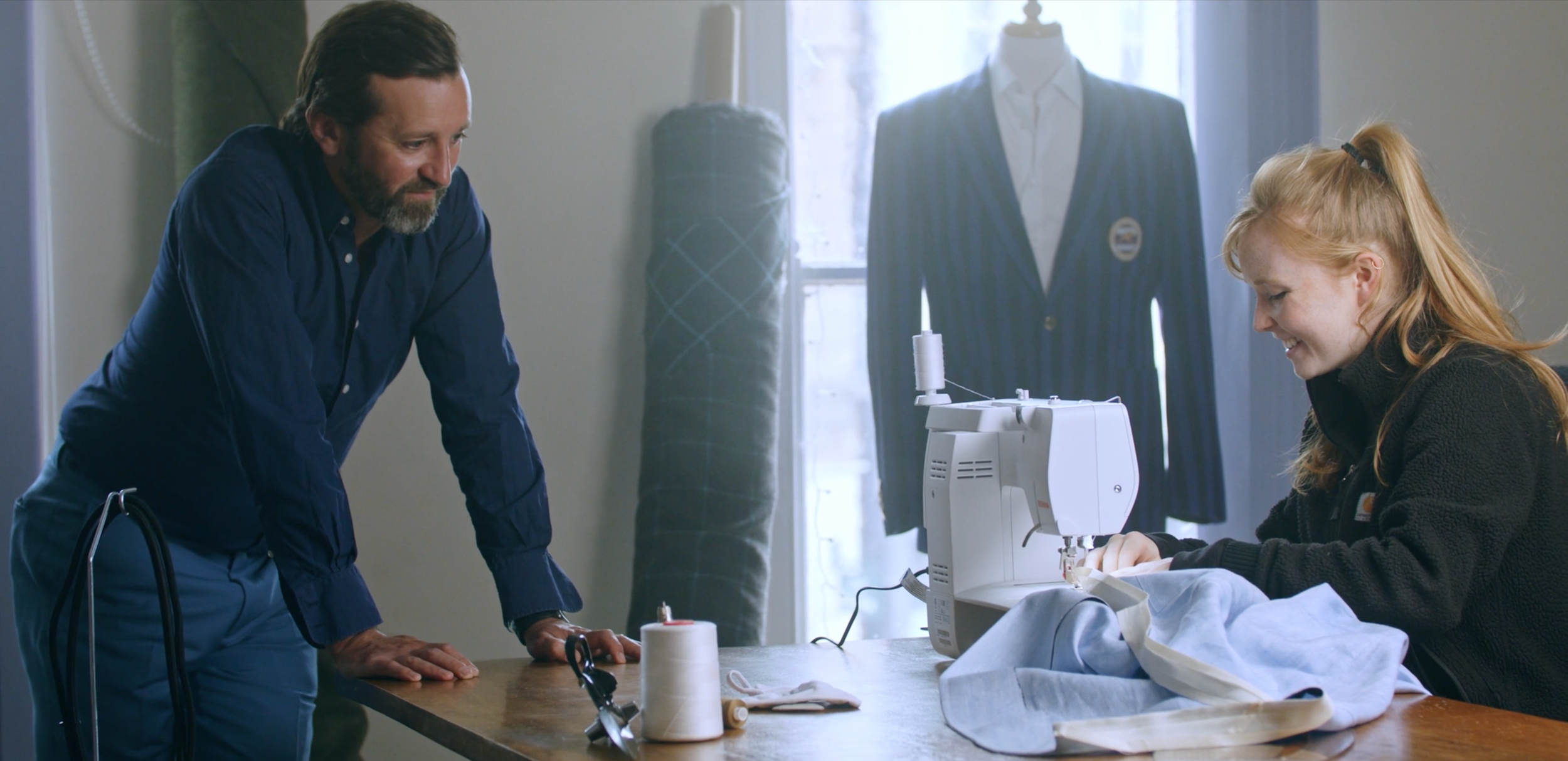A man is leaning over a sewing table, watching a red-haired woman sewing fabric using a sewing machine, in a well-lit room with a tailored suit on a mannequin and fabric rolls in the background.