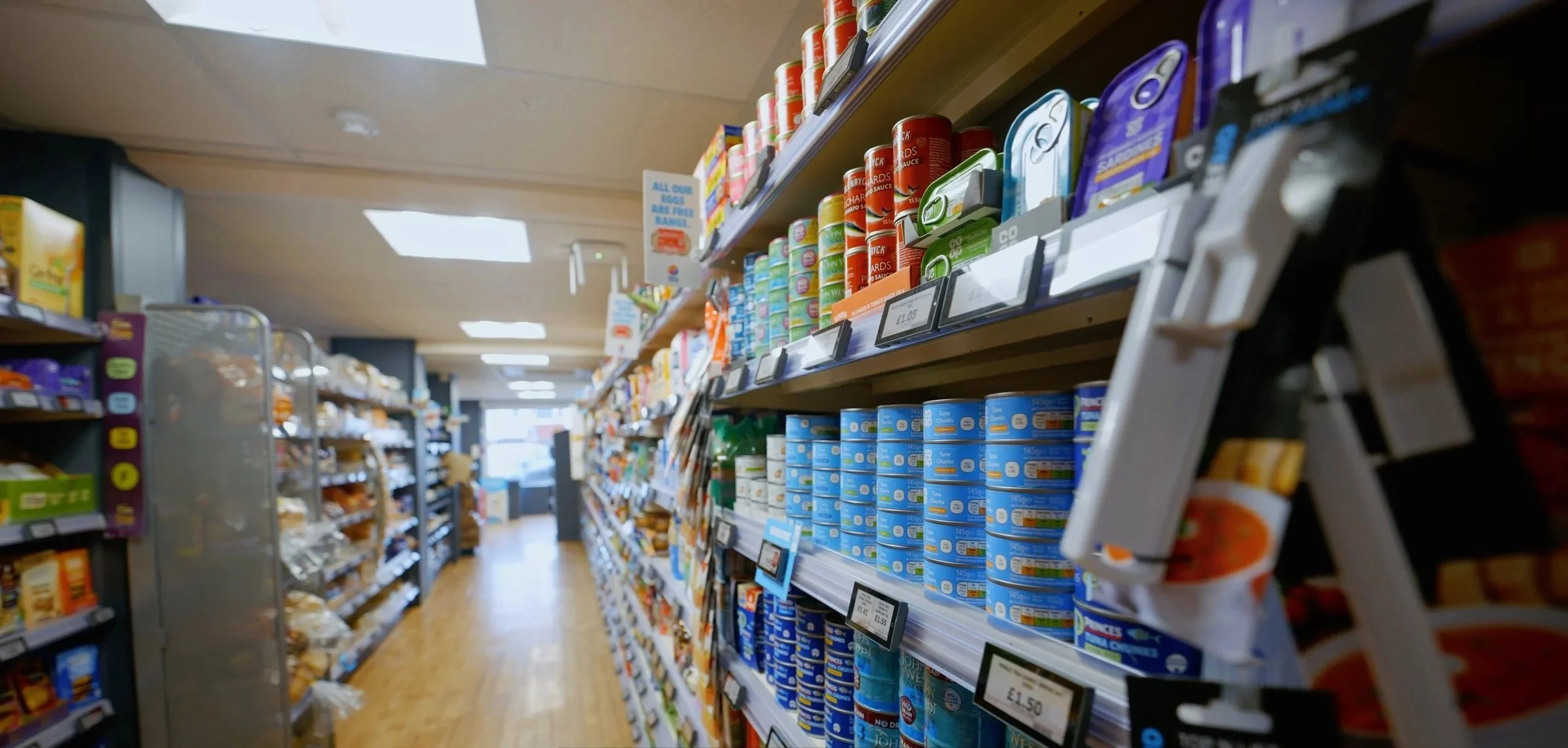 Grocery store aisle with shelves stocked with canned and packaged foods, with price tags visible. There is a step ladder partially visible on the right, and the store has wooden flooring and bright lighting.