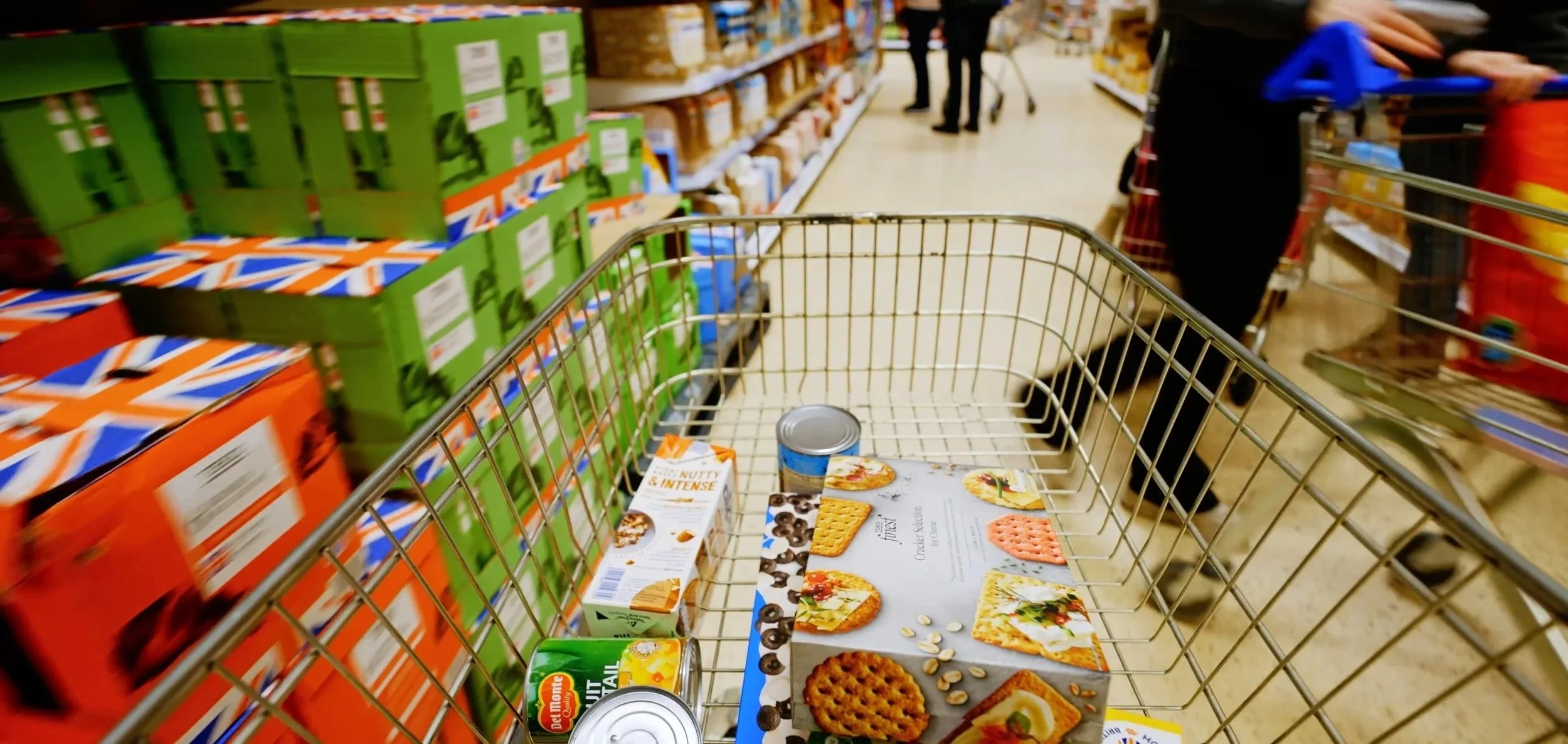 A shopping cart in a grocery store aisle with various food items inside, including canned goods, boxes, and promotional flyers, with shelves stocked with boxes and packages on either side.
