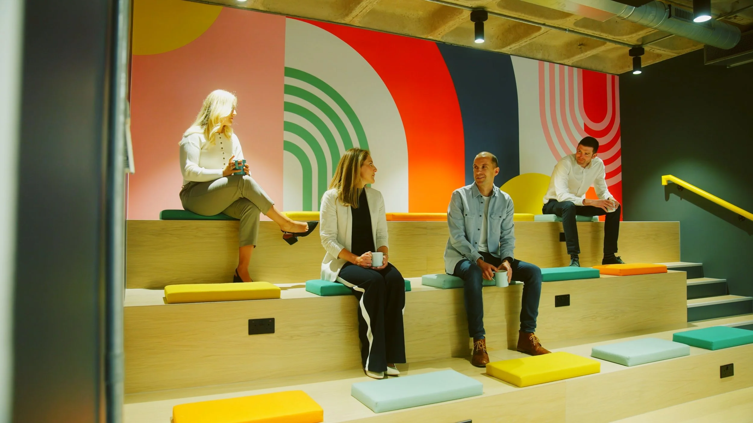 Four people sitting on multicolored stepped seating in an office or modern interior space, with a vibrant, geometric wall art in the background and ceiling lights.