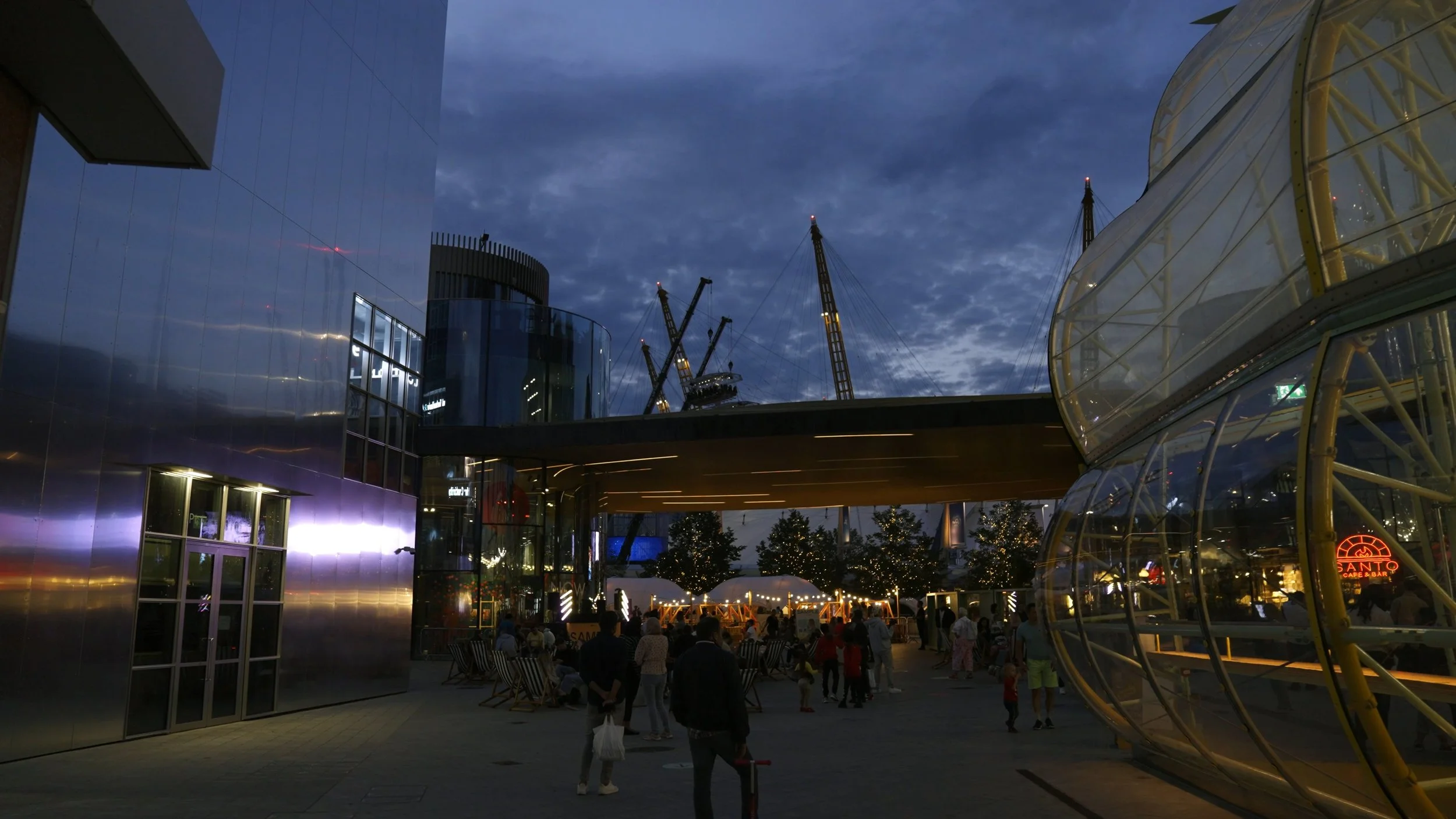 Nighttime scene at an outdoor shopping or entertainment complex with modern glass buildings, trees with lights, and a crowd of people walking around.