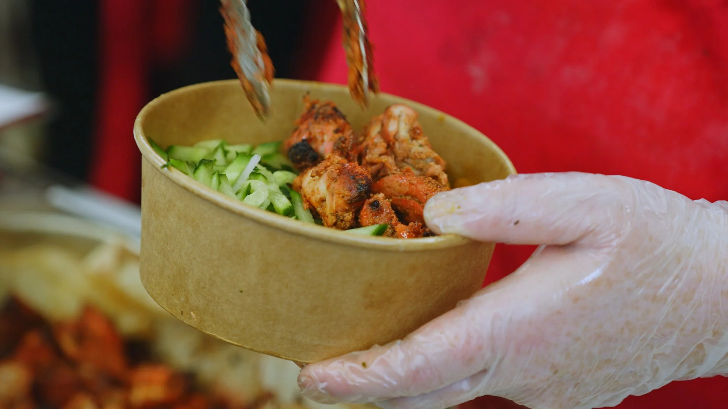 A hand wearing a disposable glove holds a brown takeout container with sliced cucumbers and grilled chicken pieces.