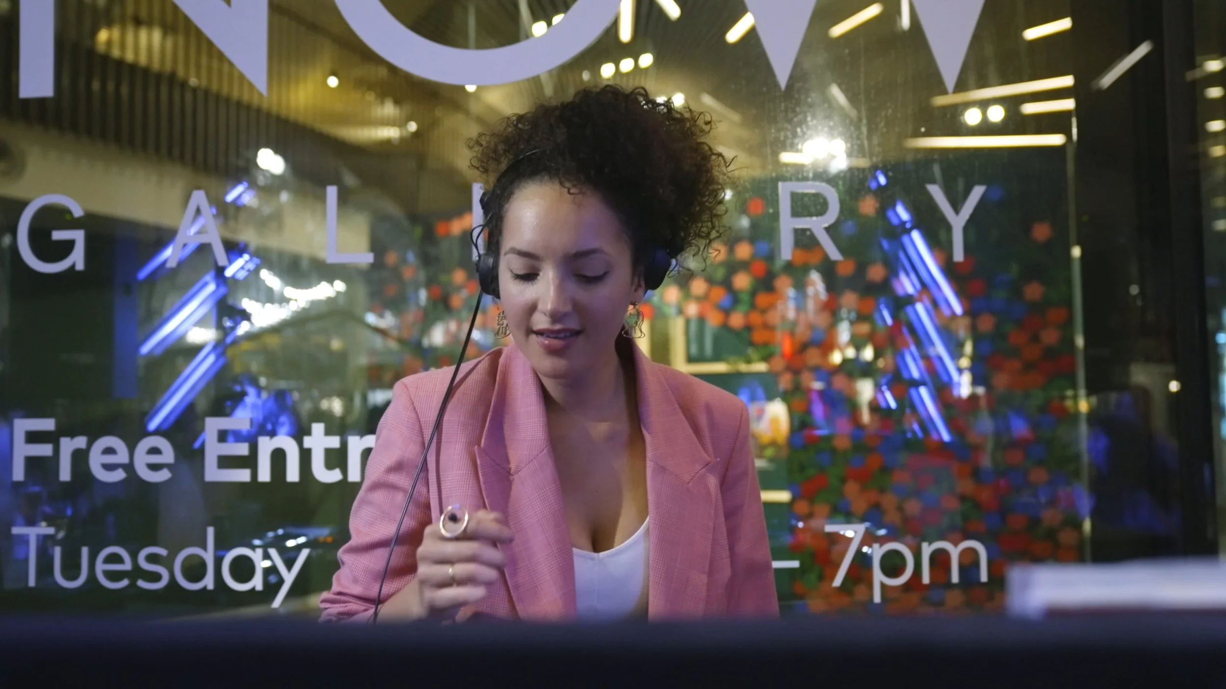 A woman wearing headphones and a pink blazer looking down, seen through a glass door with text advertising a gallery, free entry, and opening hours.