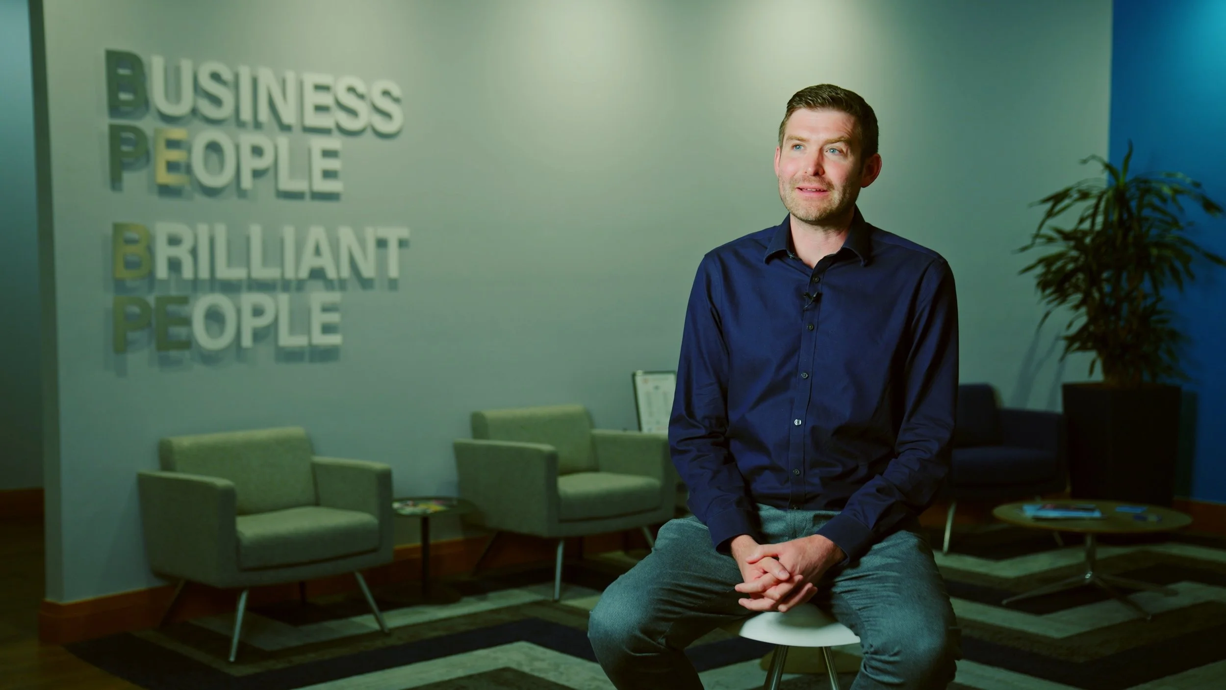 A man in a dark blue shirt sitting on a white stool in an office lounge area with green chairs, a coffee table with magazines, a large potted plant, and a wall with the words 'Business People Brilliant People' in the background.