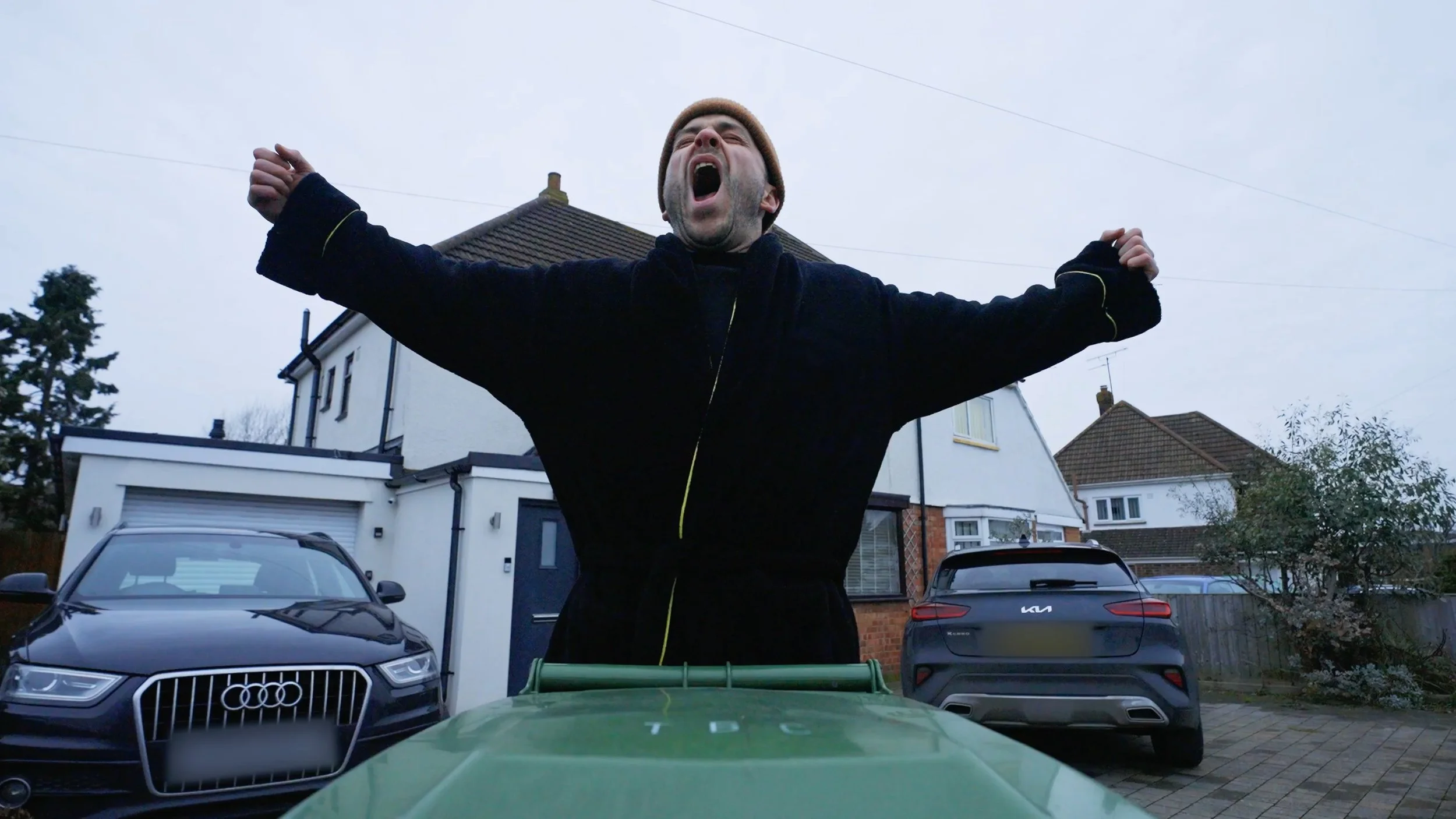 A man with arms raised and mouth open standing in a driveway in front of parked cars and houses on a cloudy day.