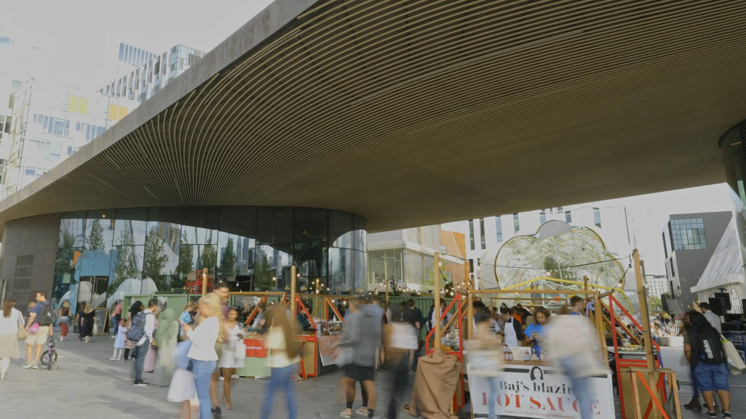 Outdoor marketplace with vendor stalls and a crowd of people in front of a modern building with a large overhang