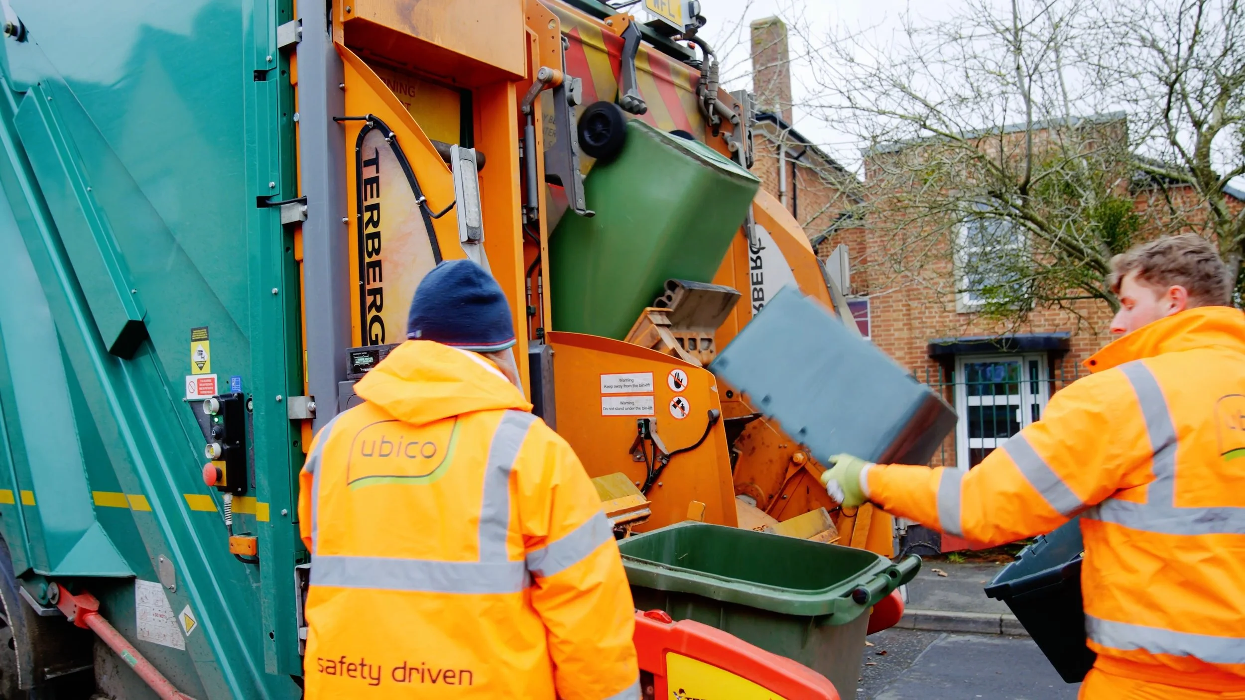 Two sanitation workers in orange reflective jackets operate a trash compacting truck on a residential street with brick houses and leafless trees in the background.