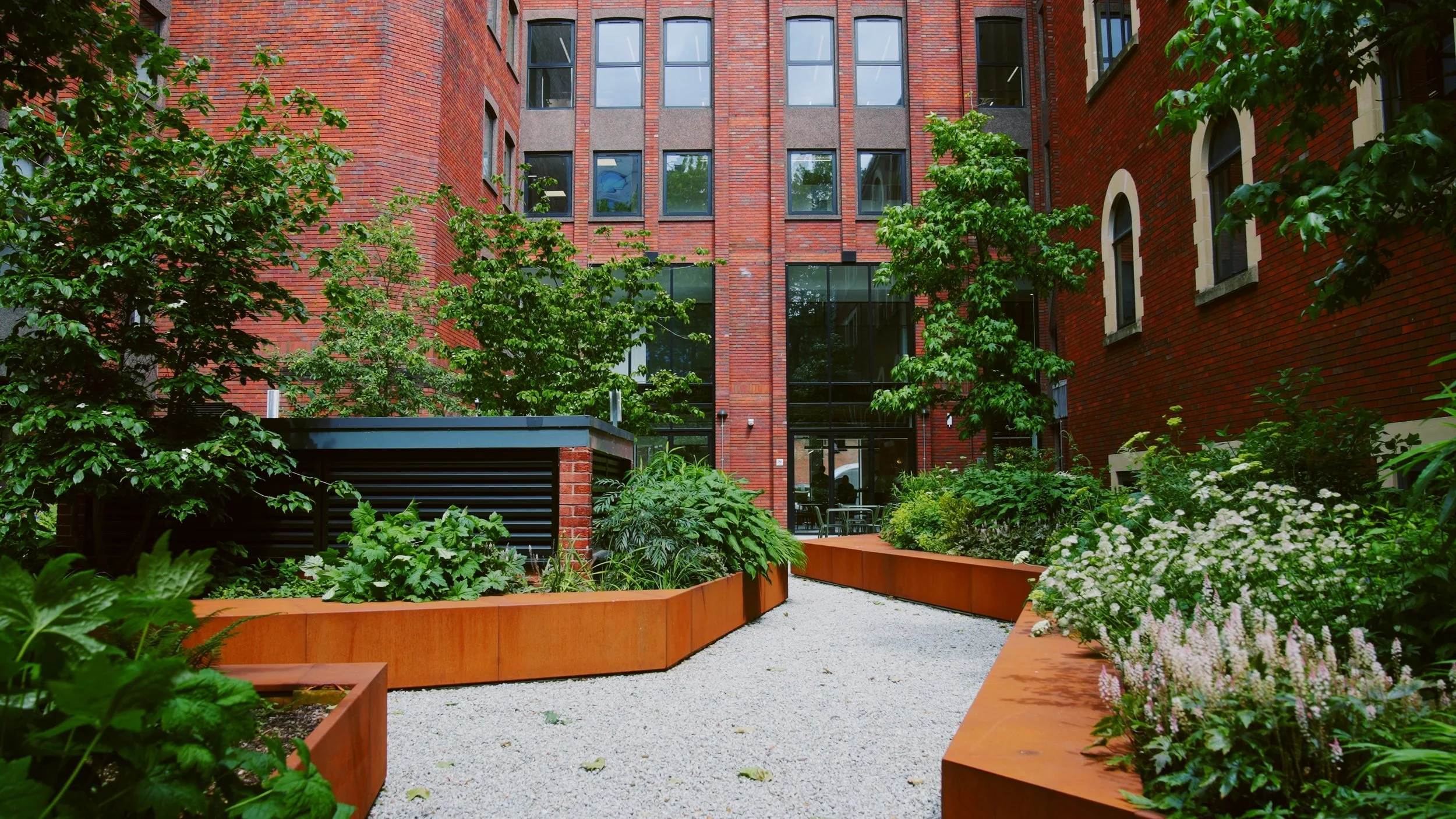 A modern urban courtyard with brick buildings, lush green trees, and landscaped plants in rust-colored planters, leading to a glass entrance.