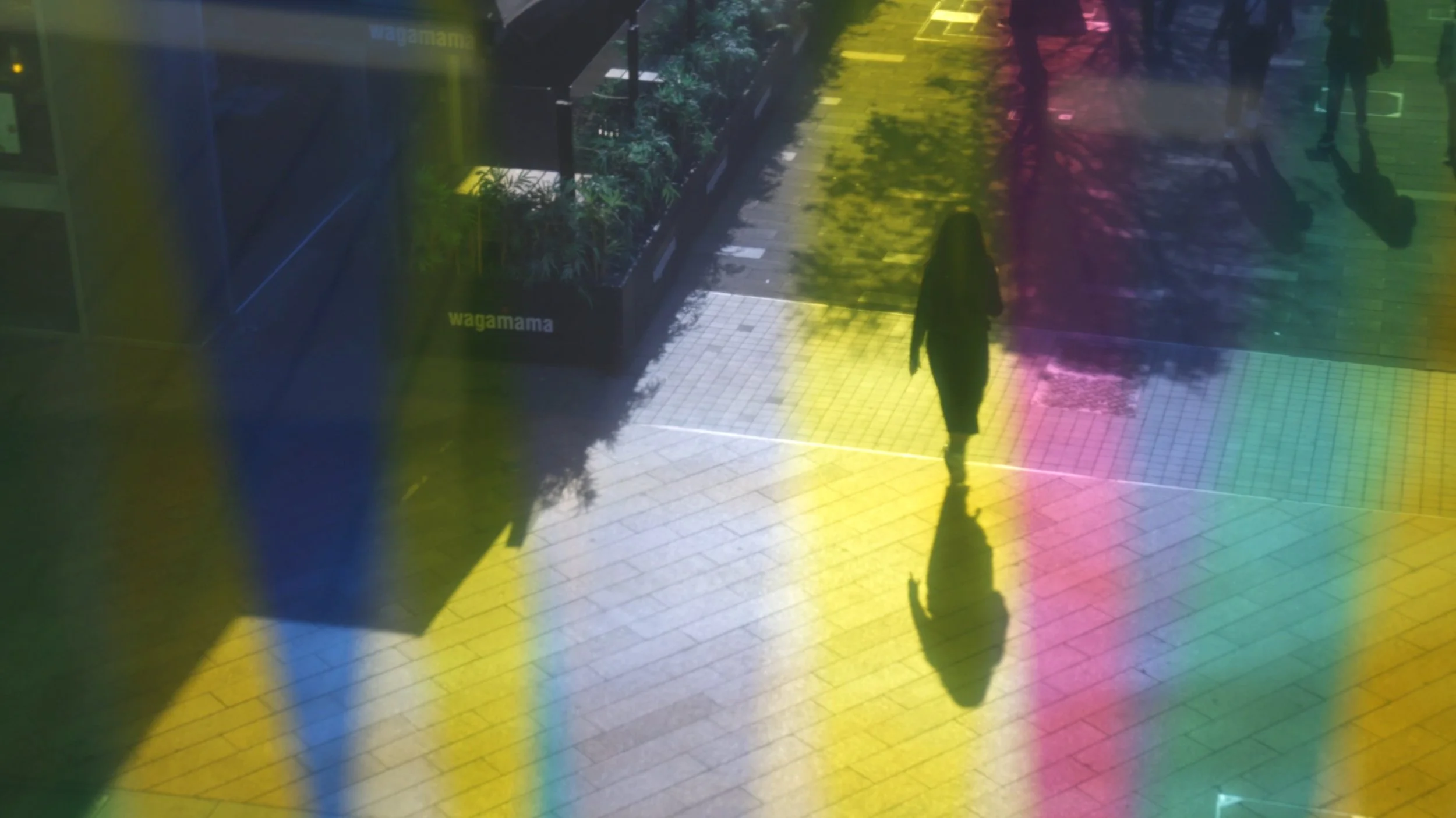 A person walking on a city sidewalk with colorful reflections and a shop window displaying the word 'wagamama.'