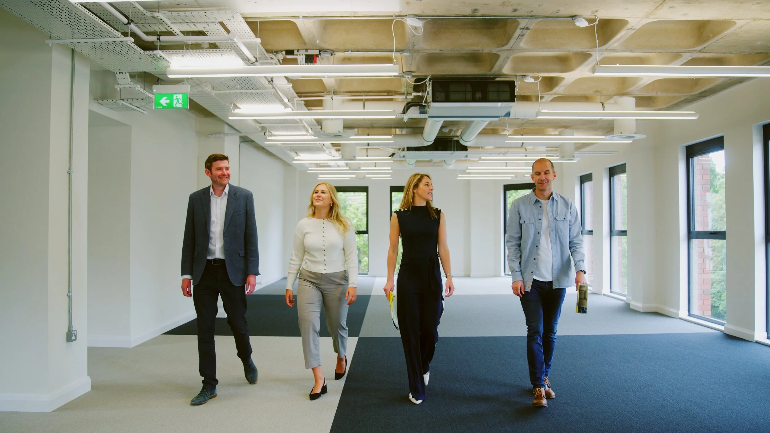 Five people walking and talking in a modern office space with large windows, bright lighting, and industrial-style ceiling.