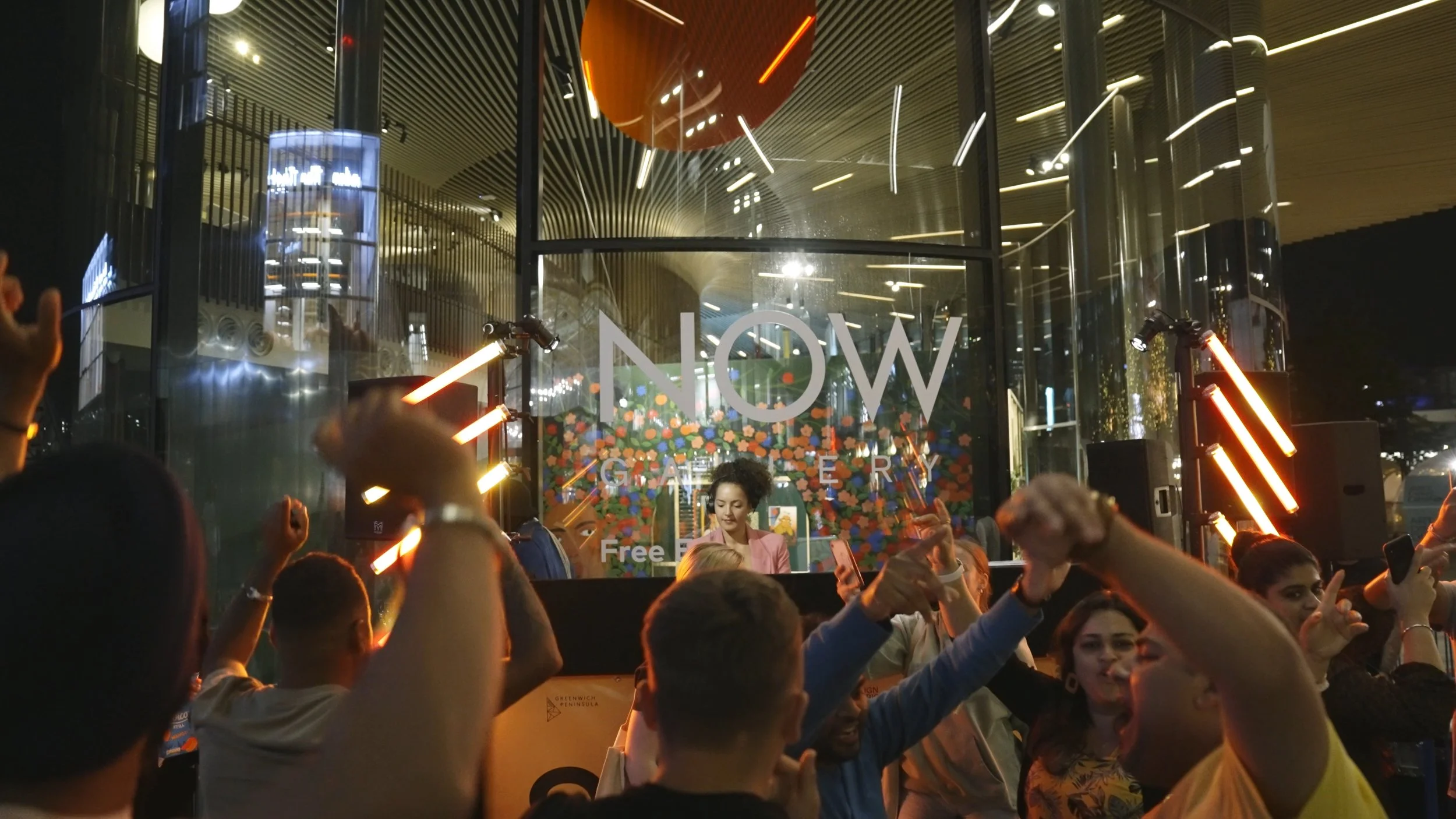 People dancing and celebrating outside an art gallery at night, with a large glass window displaying the words 'NOW GALLERY' and colorful artwork inside.