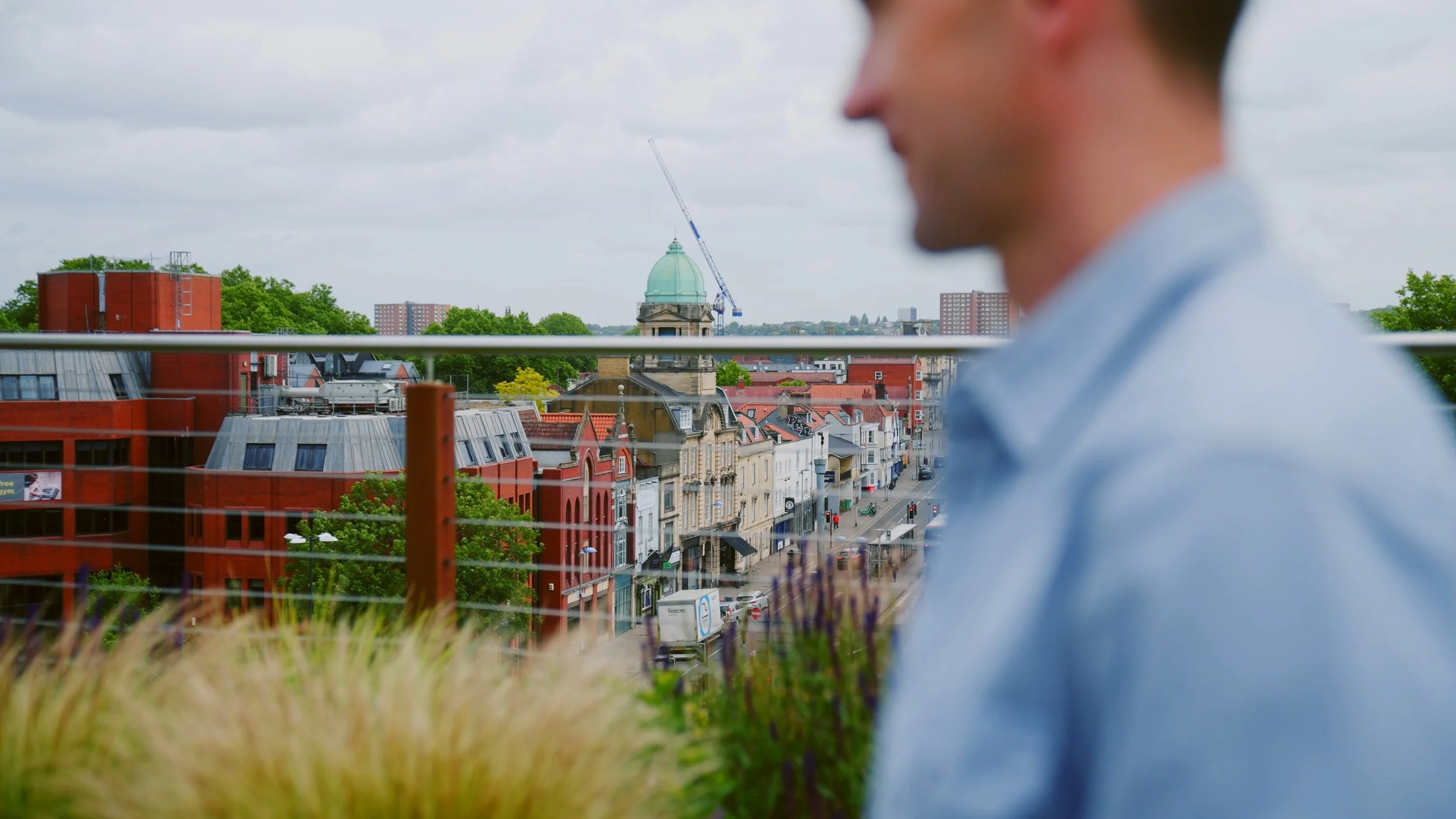 Partially blurred man in light blue shirt in foreground overlooking a city street with historic buildings, trees, and a church with a green dome, under cloudy sky.