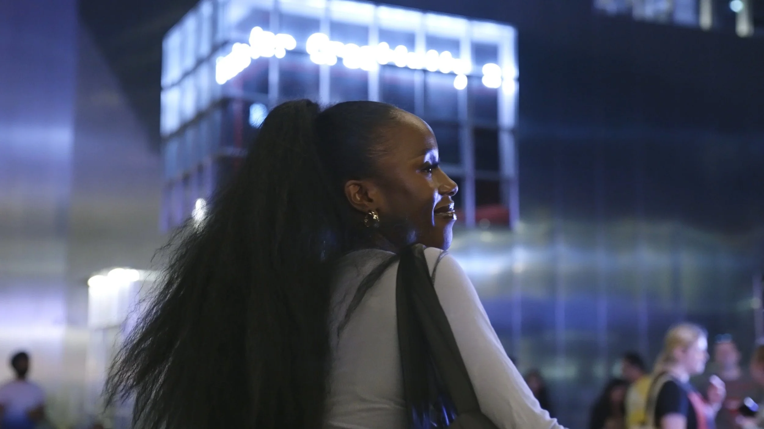 A woman with long black hair smiling as she looks to her right at night, with a modern illuminated building in the background and people around.