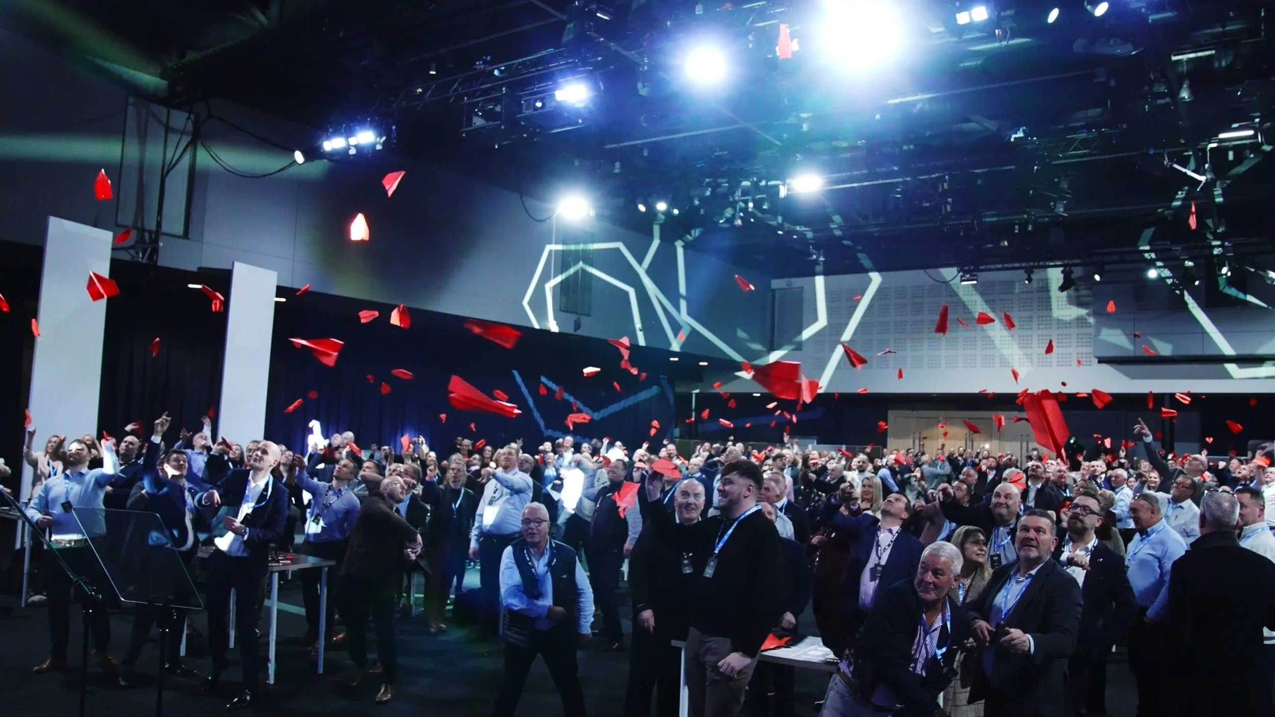 A large crowd of people at a conference or event, releasing red paper airplanes into the air in a spacious, modern indoor venue with high ceilings and bright lighting.