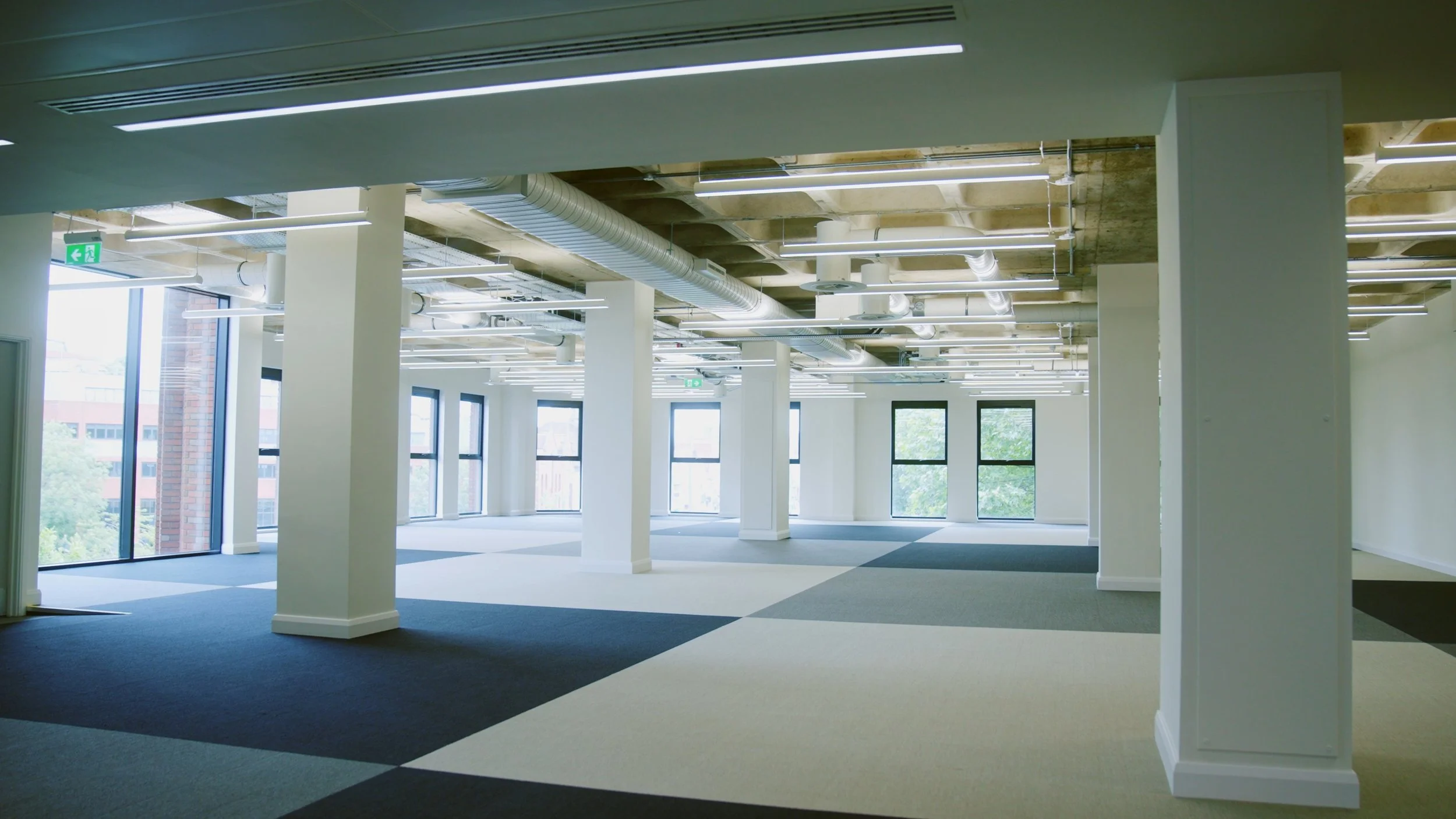 Empty modern office space with large windows, white pillars, and exposed ceiling ducts and pipes, illuminated by natural light.