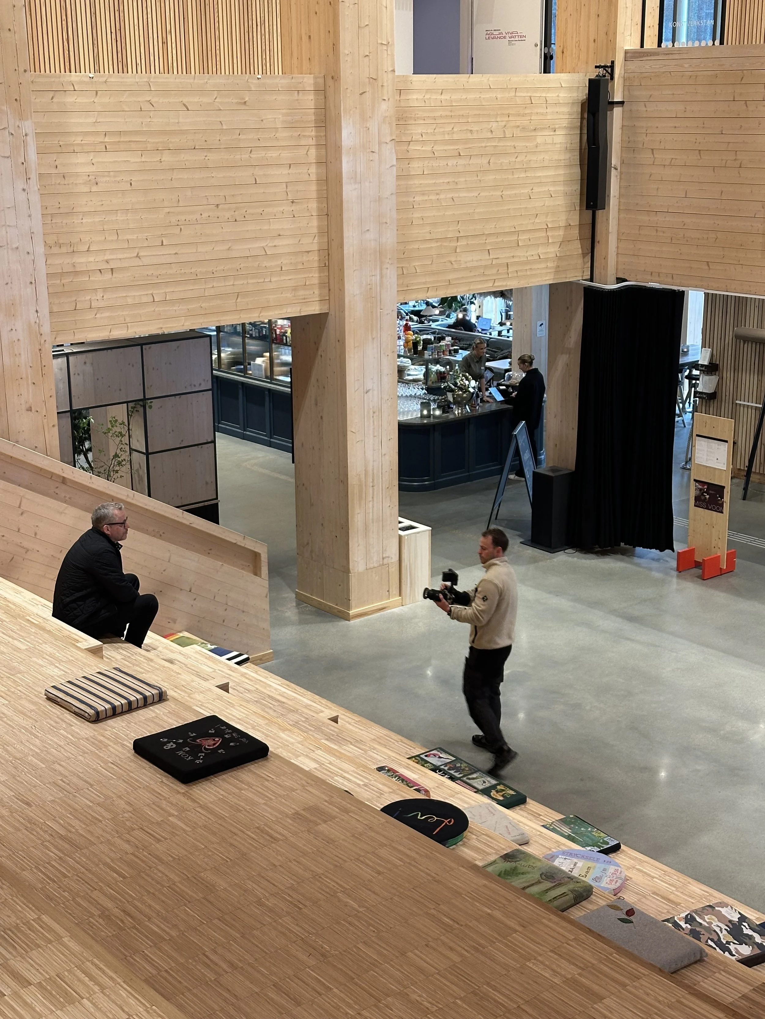 A man holding a camera walking in a modern, wood-finished cafe or restaurant with a barista counter in the background, and another man sitting on a wooden staircase.