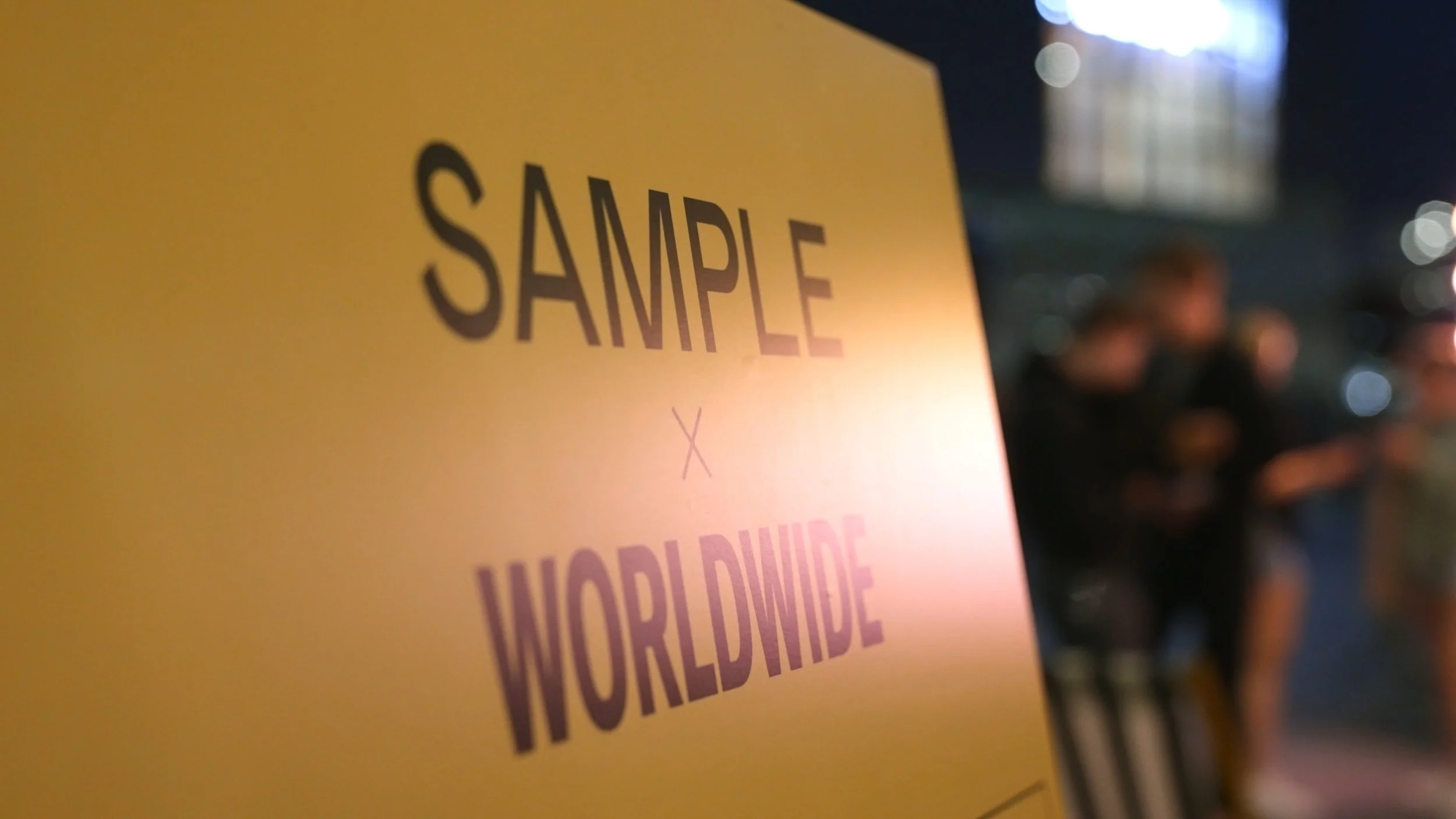 Close-up of a yellow sign with the words 'SAMPLE' and 'XX WORLDWIDE' visible, with blurred figures of people in the background at night.