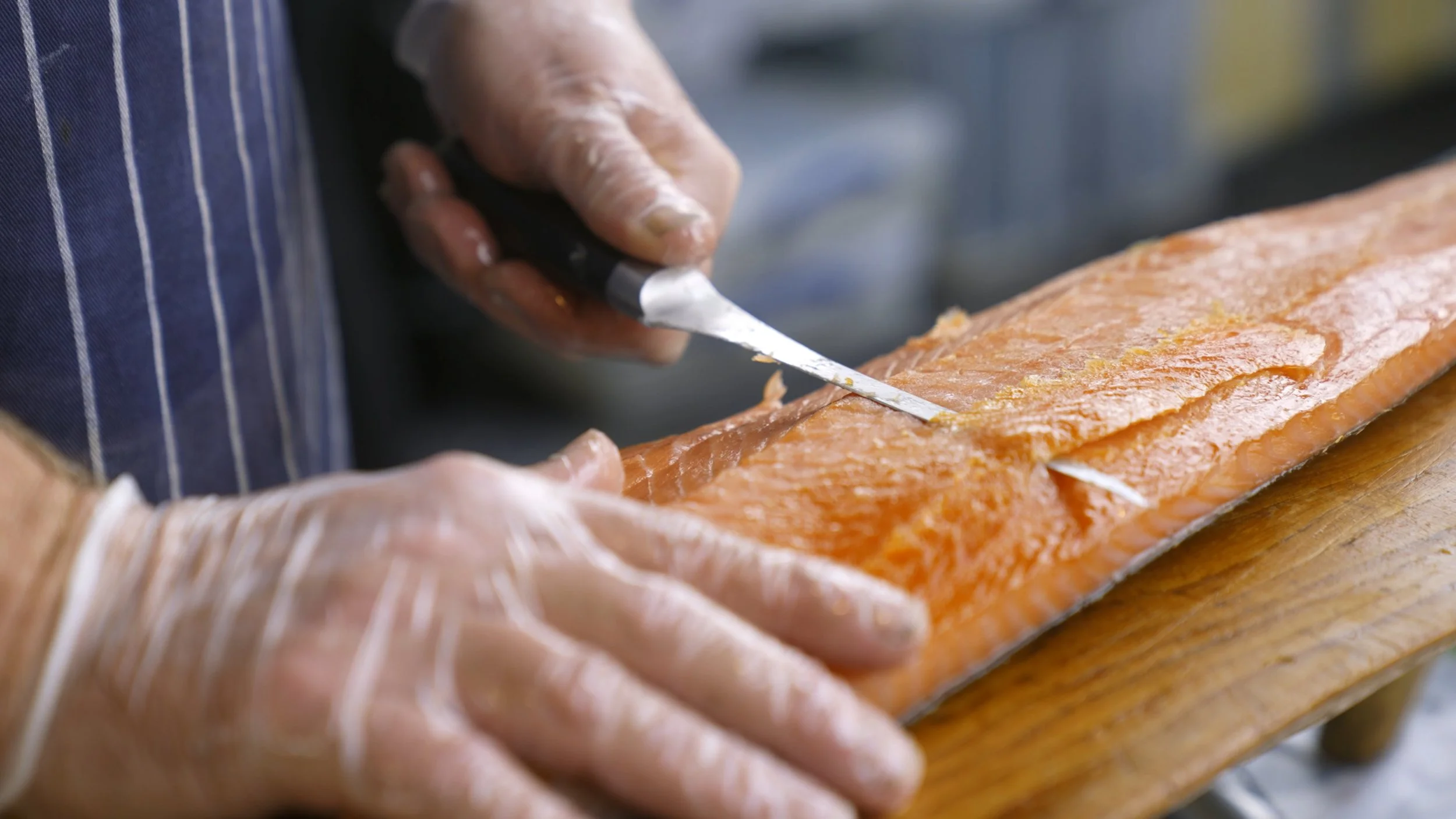 Close-up of a person wearing gloves and cutting a fresh salmon fillet on a wooden cutting board in a kitchen.