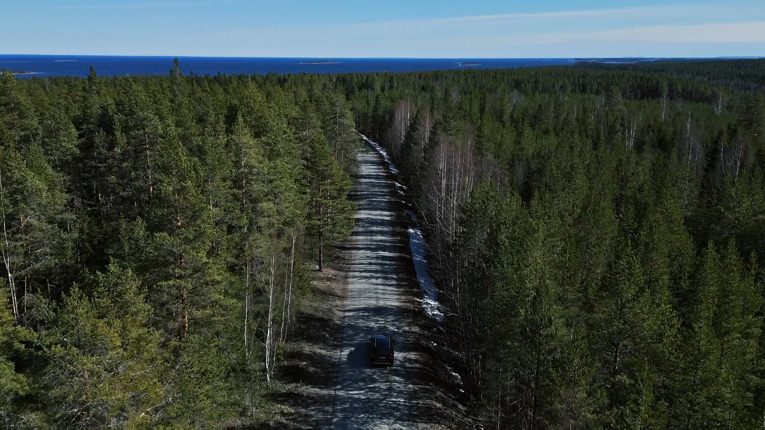 An aerial view of a car driving on a narrow dirt road through a dense forest with a distant view of a lake and blue sky.