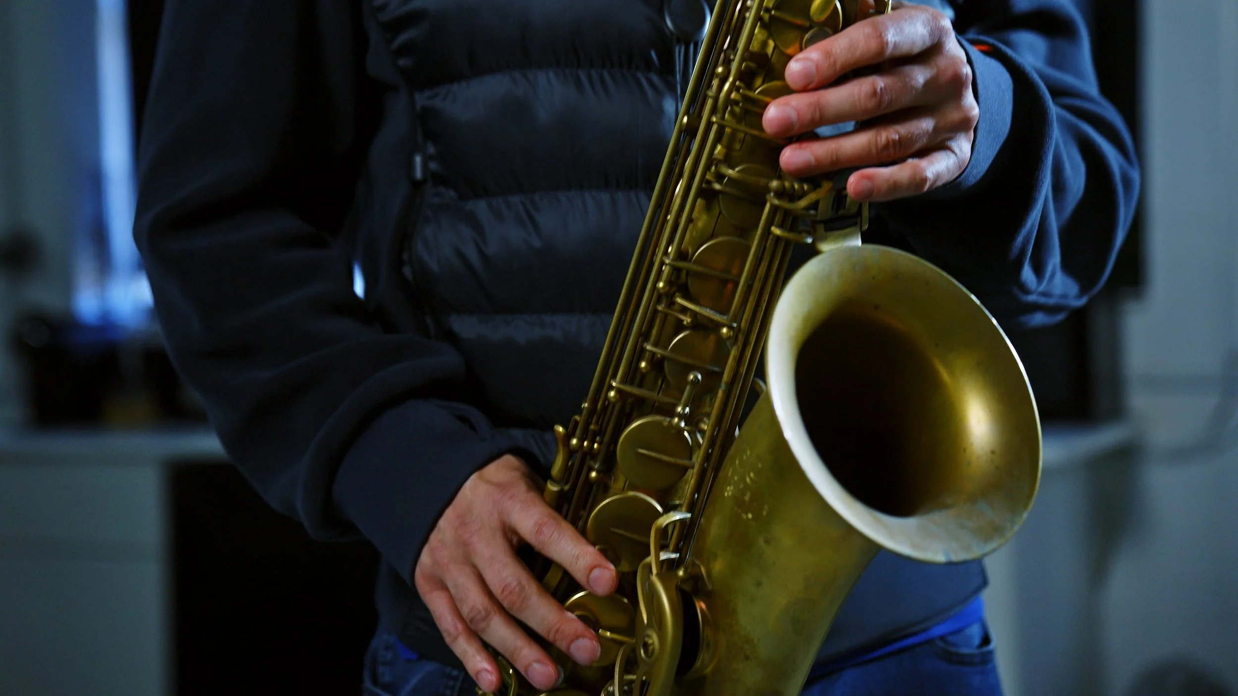 Close-up of a person playing a brass saxophone, wearing a black jacket, in an indoor setting.