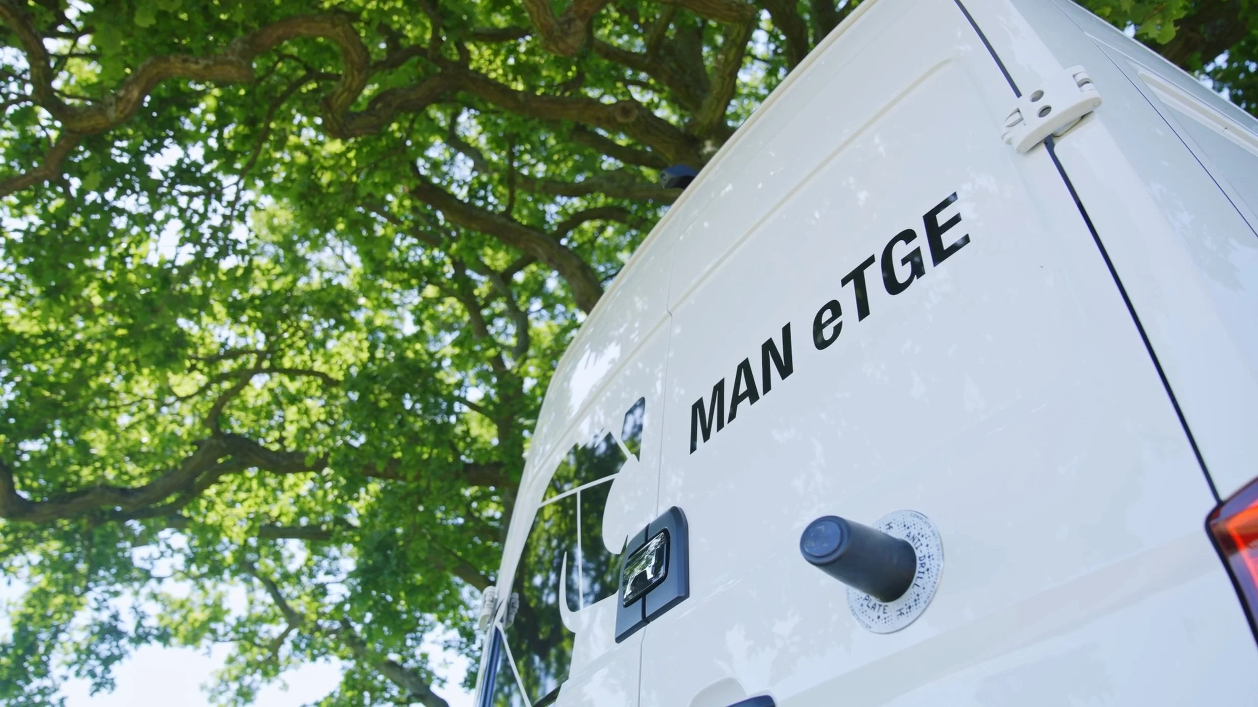 The back of a white emergency vehicle with the words "MAN e TAGE" on it, parked underneath a green leafy tree.