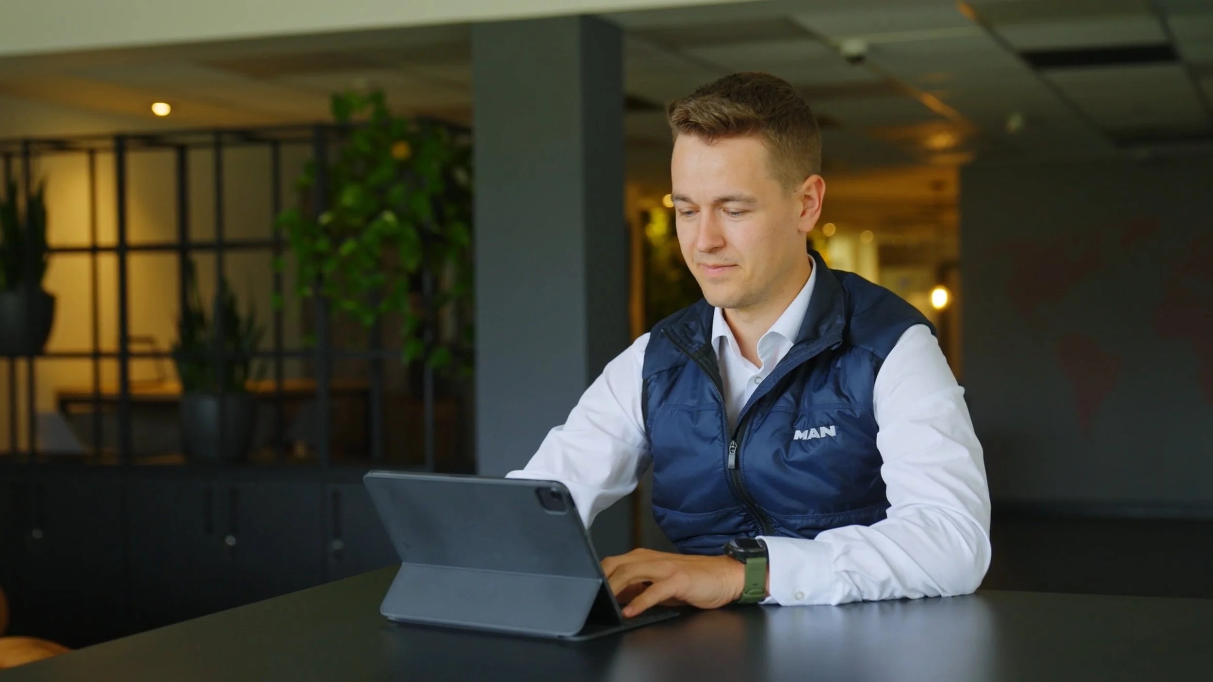 A young man with short light brown hair and a slight smile, wearing a white shirt and blue vest, is sitting at a black table in a modern indoor setting, using a gray tablet with a foldable cover. There are green plants and decorative lighting in the